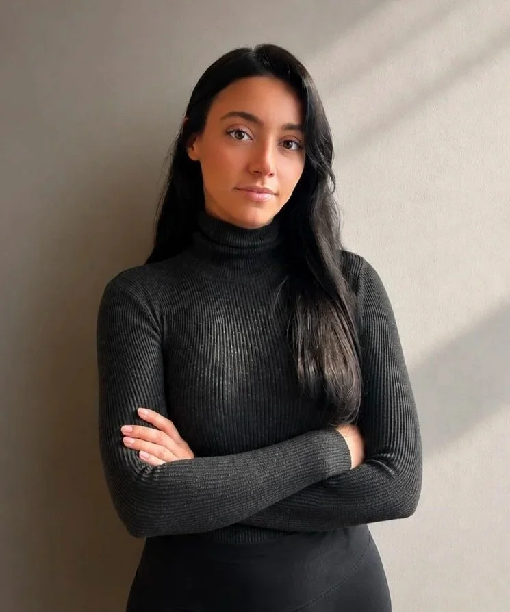 A young woman with long dark hair, wearing a black ribbed turtleneck, standing with her arms crossed against a plain beige wall.