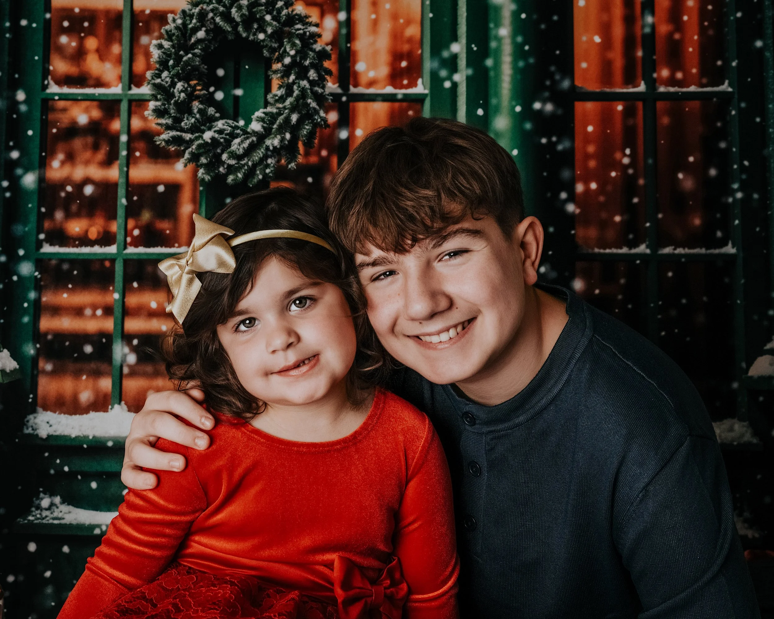 A young girl and a teenage boy posing closely, with a snowy Christmas wreath on a window and snowflakes falling in the background.