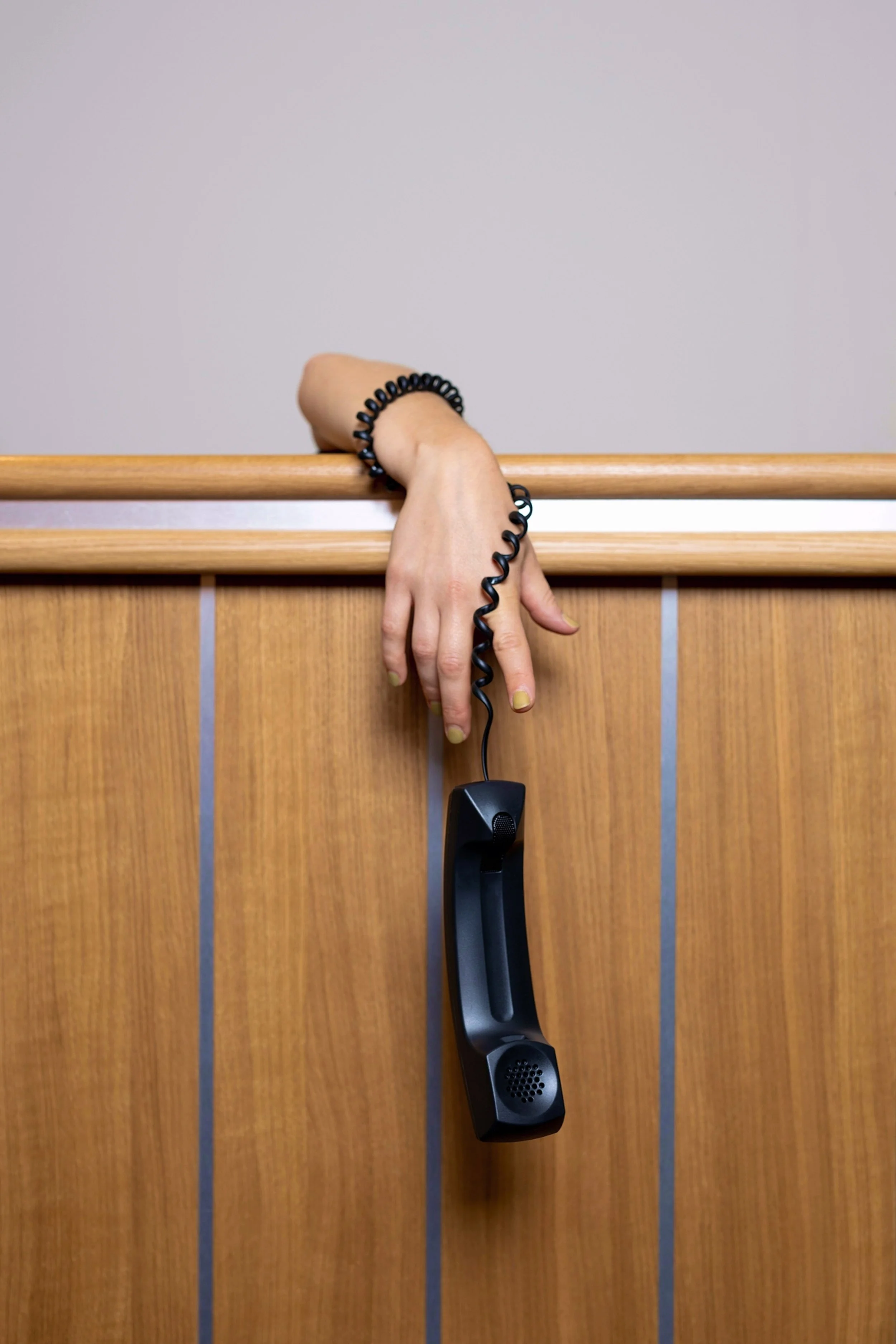 A person’s hand with a black spiral cord, a black bracelet, and painted fingernails resting on a wooden railing with a vintage black phone hanging from the cord.