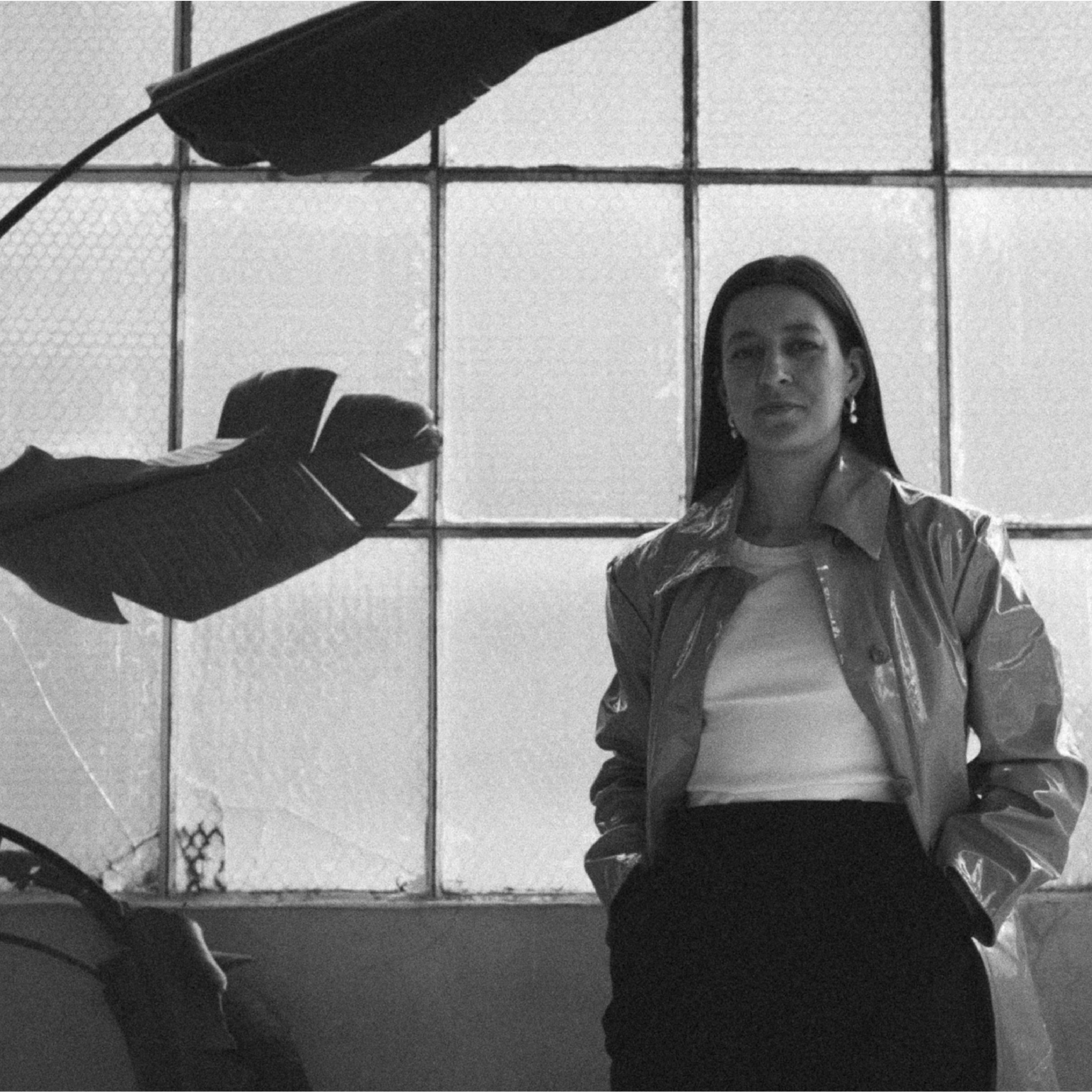 A woman with dark hair, wearing earrings, a shiny jacket, and a white top, standing with hands in her pockets in front of a window grid, with a large leaf in the foreground.