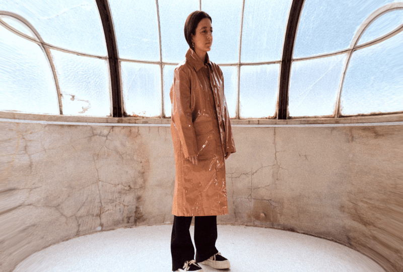 A woman wearing a brown raincoat and black pants standing in an arched glass structure with cracked walls.