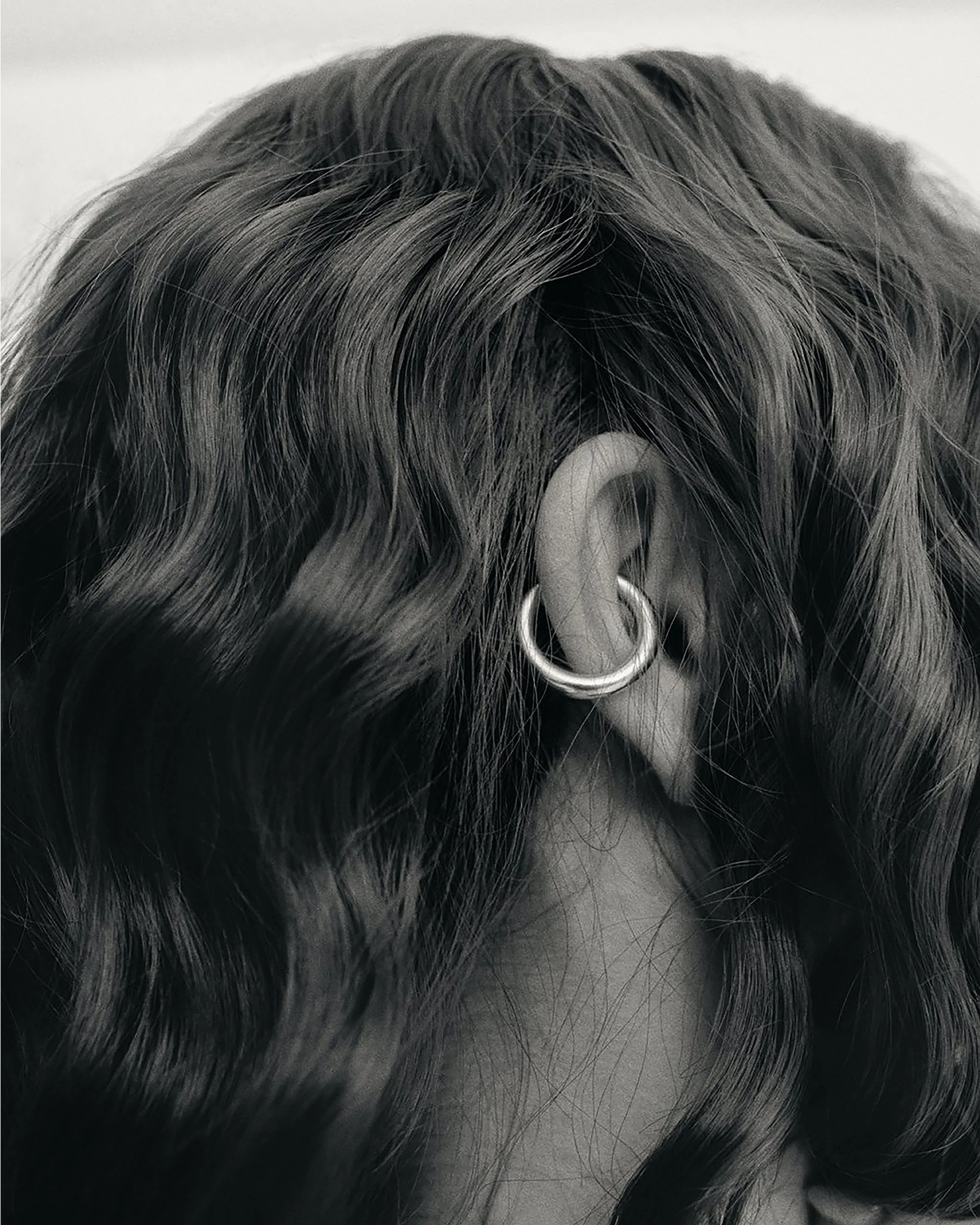 Close-up of a person's ear with a hoop earring, surrounded by wavy, dark hair.