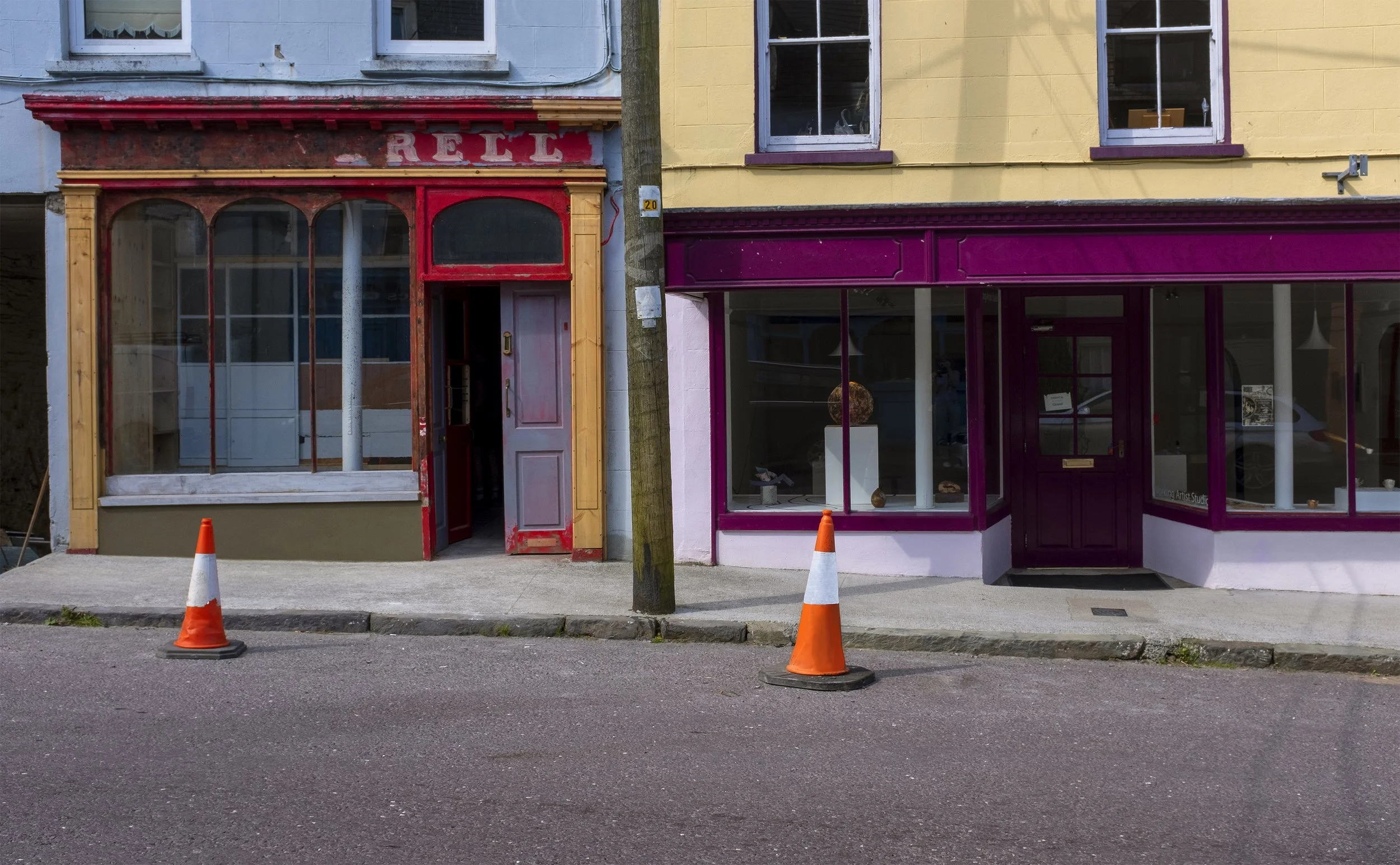 Two storefronts on a city street, one painted light blue with a red and yellow trim and the other painted yellow with purple accents. There are two orange traffic cones on the sidewalk in front of the shops, and a utility pole between the buildings.