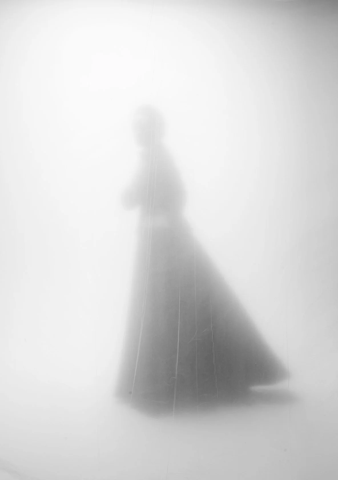 A person in a long coat or cape, standing in front of a foggy, white background, with their back to the camera. The image is black and white and blurry, creating an abstract silhouette.