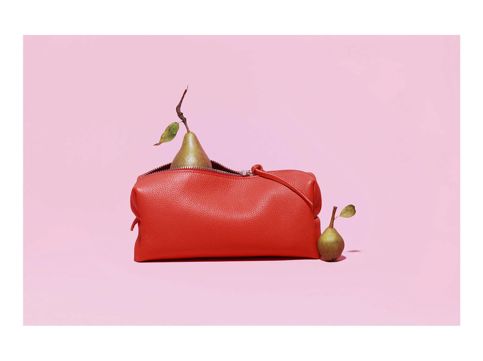 Red leather bag with two pears, one inside and one on the outside, on a pink background.