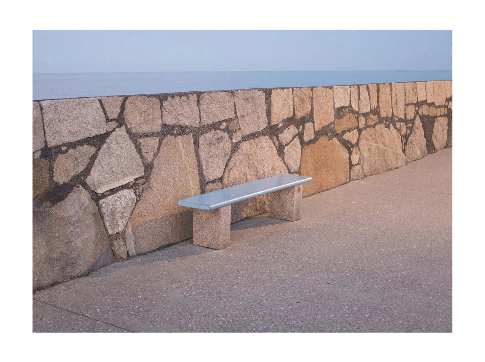 A stone wall with a concrete bench in front of it, overlooking the ocean during sunset.
