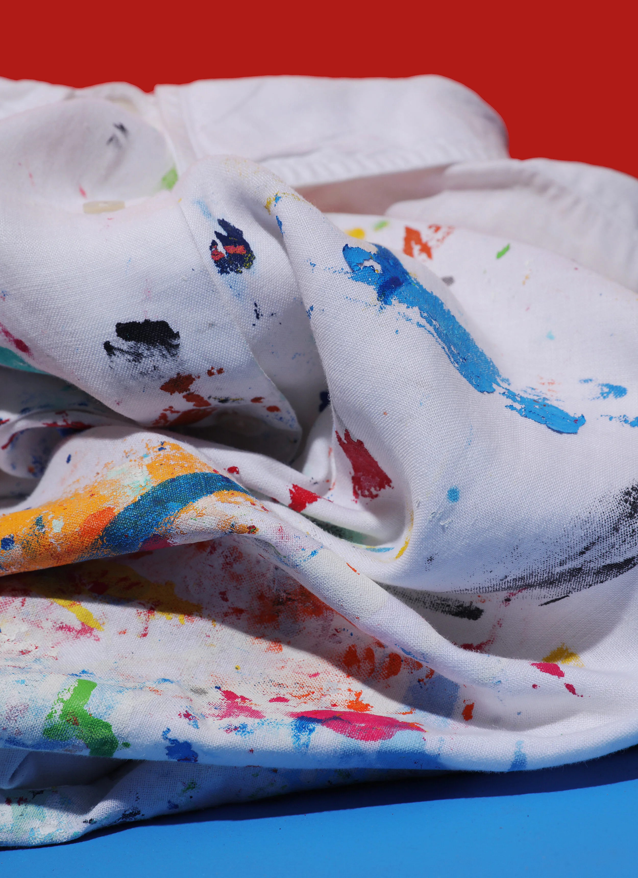 Cloth with multicolored paint splatters and smudges, crumpled on a blue surface against a red background.