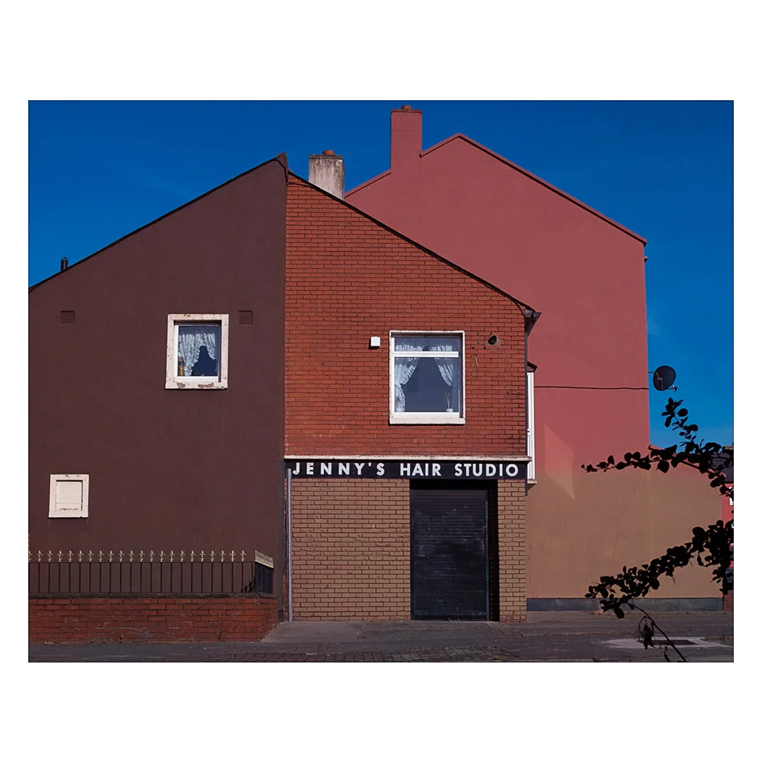 A building with a brick facade and a sign that reads "JENNY'S HAIR STUDIO." The building has two windows with lace curtains, a metal roll-up door, and is set against a clear blue sky.
