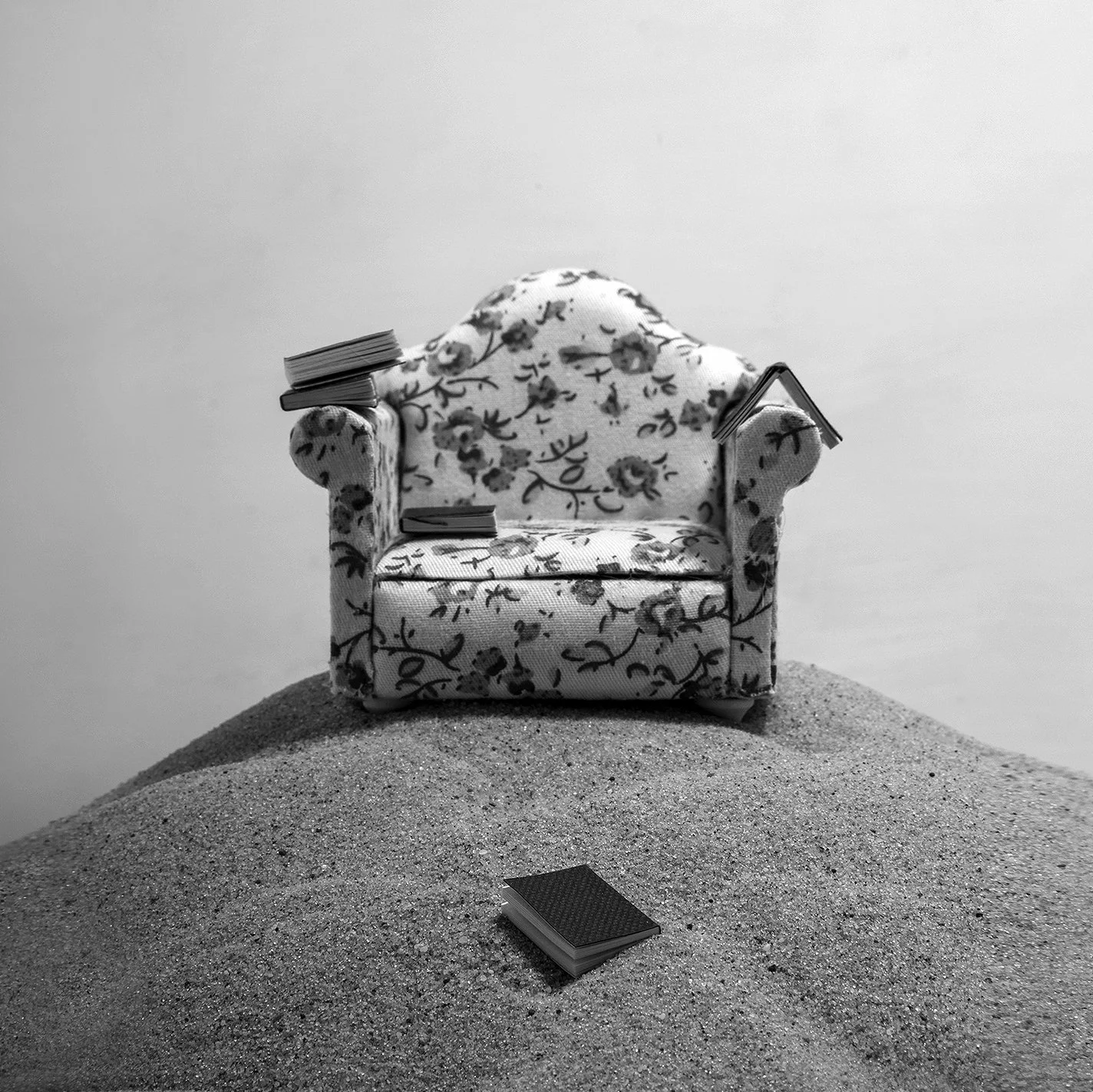 Black and white photo of a miniature floral armchair on a small mound of sand, with books  on the ground in front and small stacks of books balanced on the armchair's armrests.