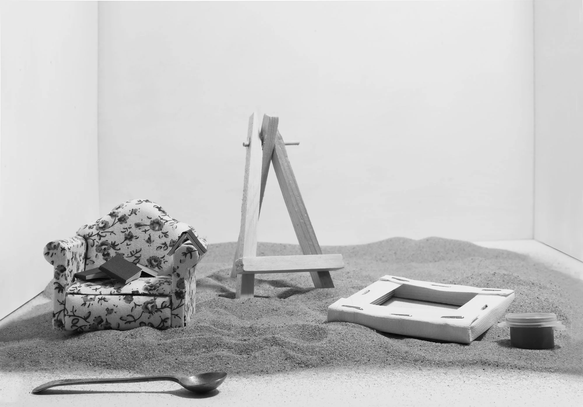 A black and white photograph of a miniature room scene with a floral-patterned armchair, a small open book on the chair, a bed made of foam or drywall with scattered papers, a small bucket, a step ladder, and a boat on the ground. The walls are plain