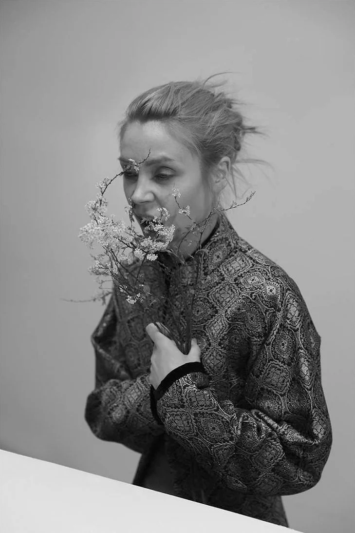A woman with messy hair holding a bouquet of flowers close to her face, smiling with her eyes closed, against a plain background.