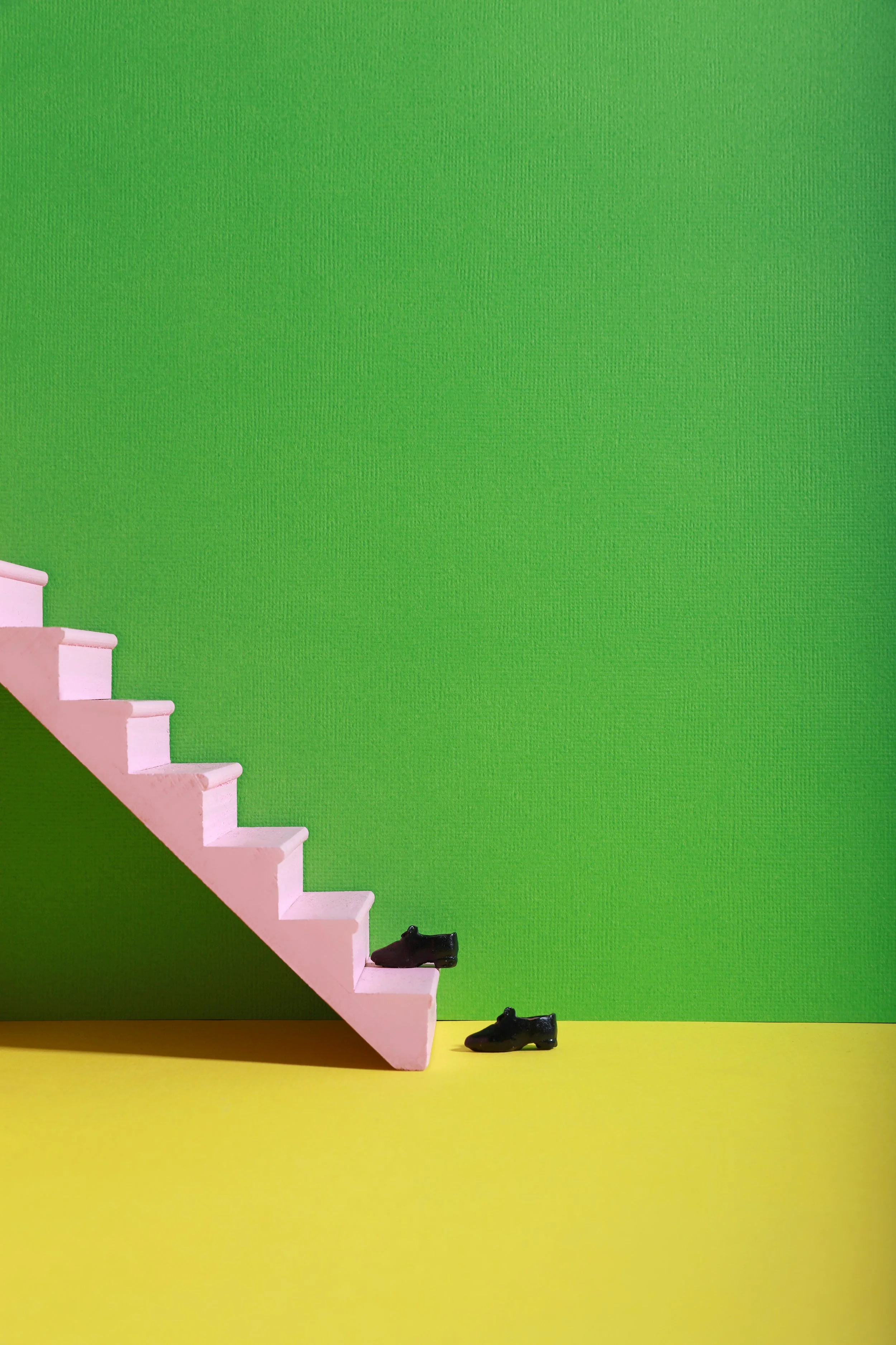 Miniature pink staircase with two tiny black shoes, set against a bright green wall and yellow floor.