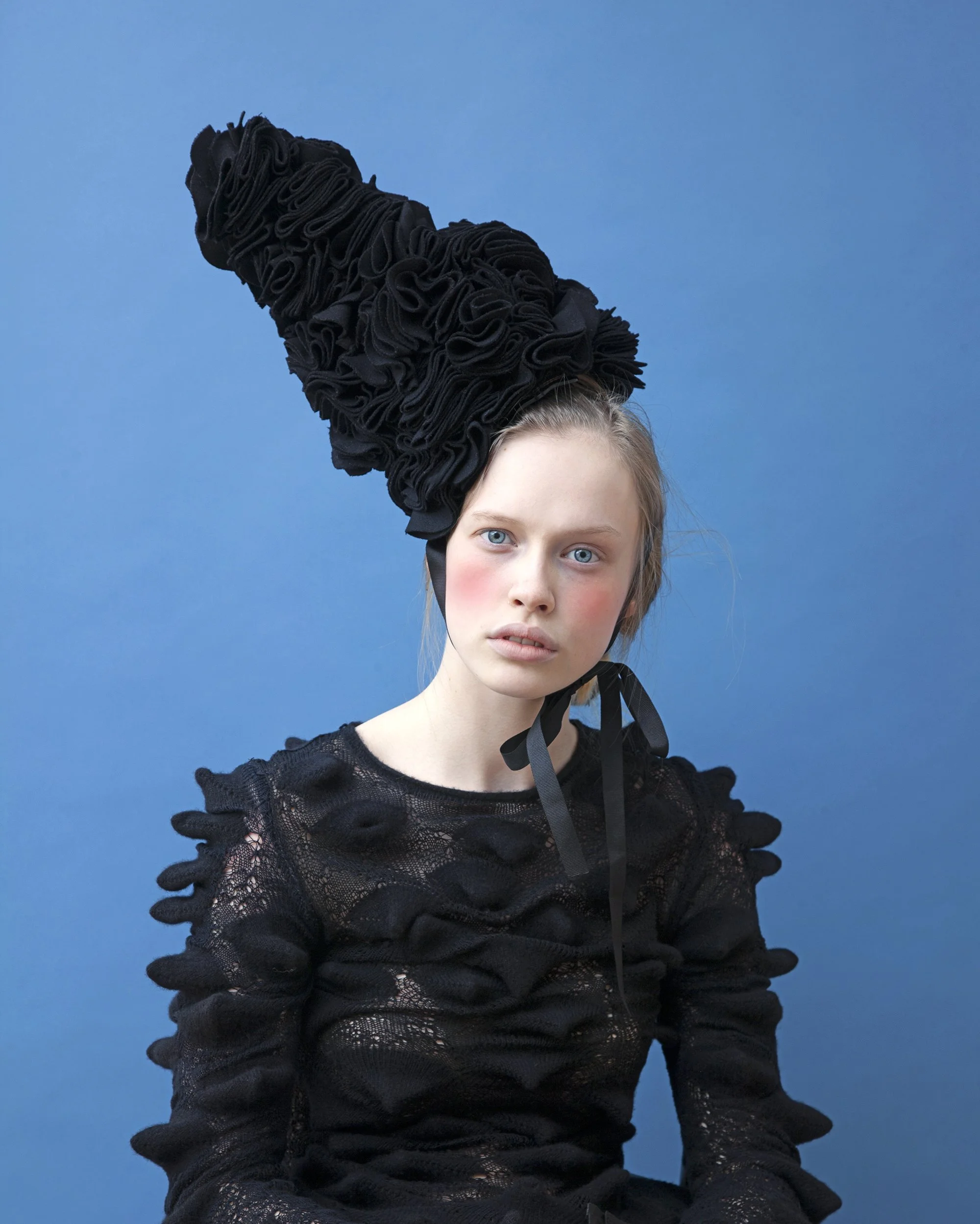 A young girl with blue eyes and fair skin wearing a black lace dress with ruffled long sleeves and a large, black, ruffled hat against a plain blue background.
