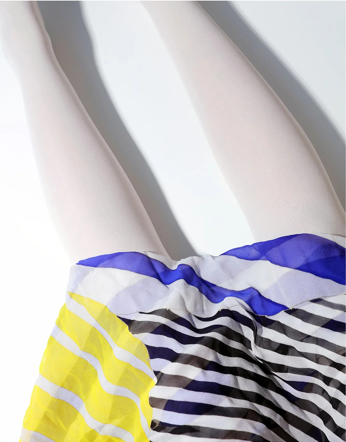Close-up of a pair of legs wearing light-colored, sheer tights, with a colorful striped elastic band or fabric around the ankles featuring yellow, black, white, and blue stripes.