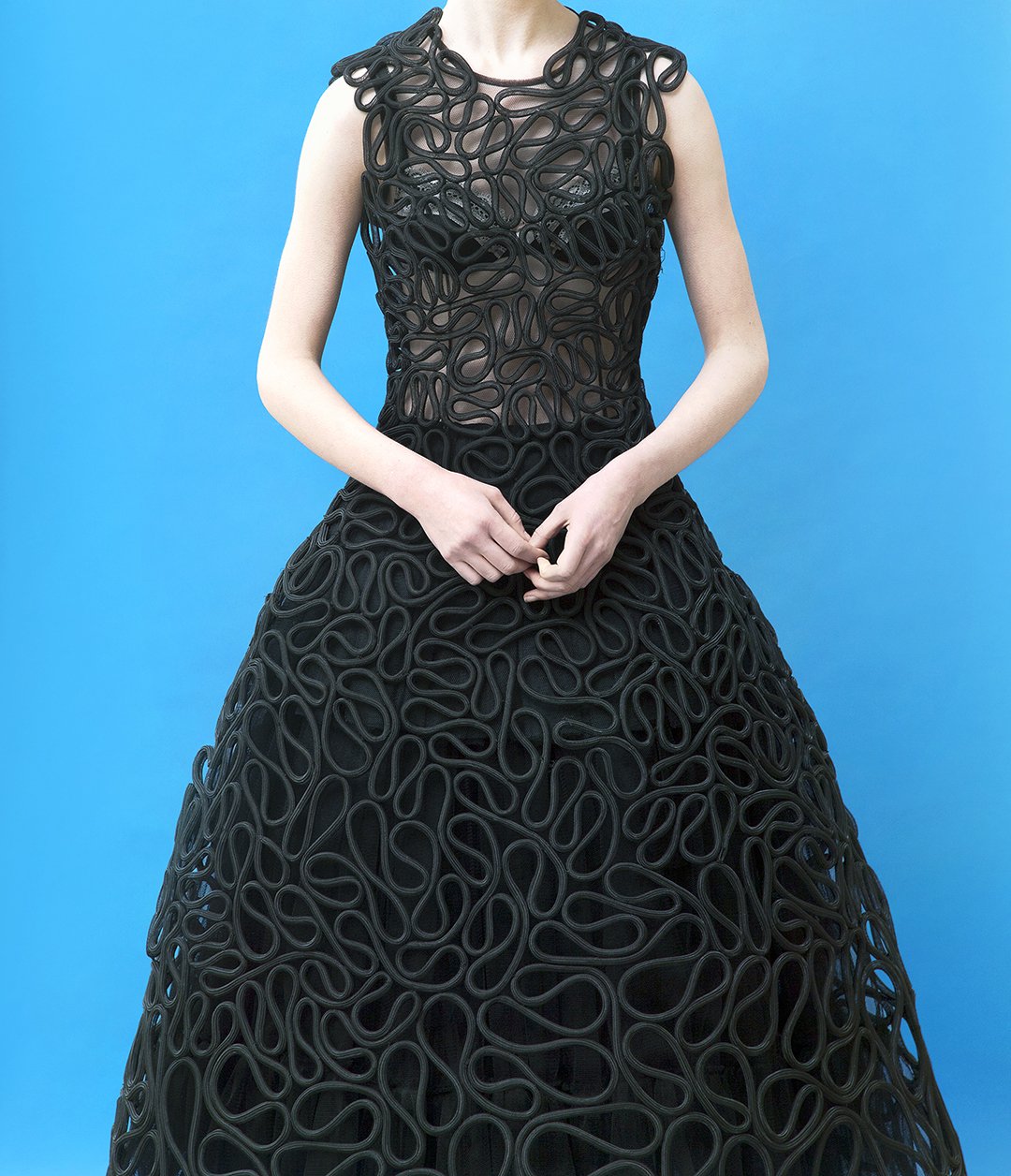 A woman wearing a black gown with intricate looped patterns against a blue background.