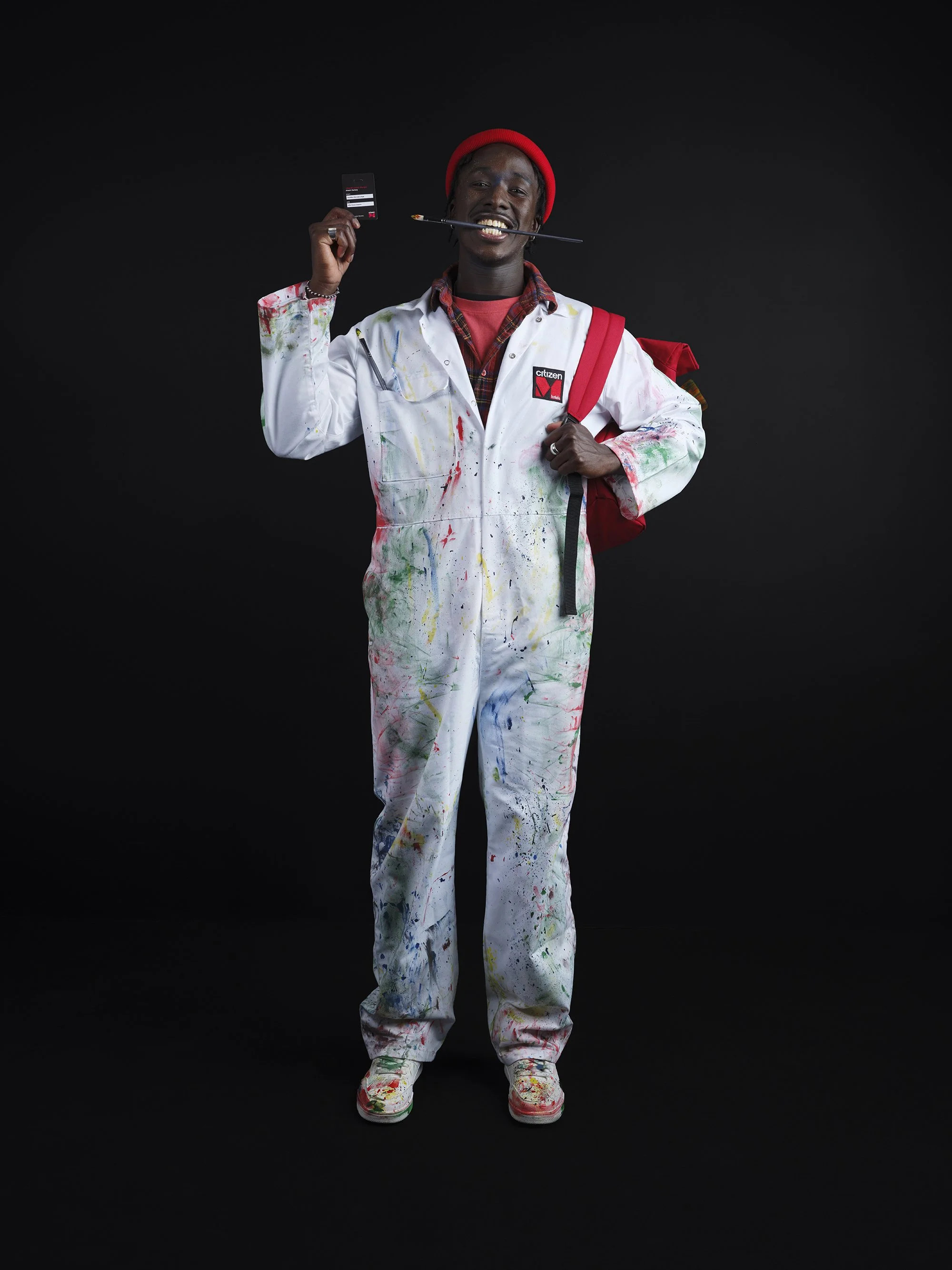 A young man wearing a red beanie and paint-splattered white jumpsuit holding a black card in his left hand and a paintbrush in his mouth, standing against a black background.