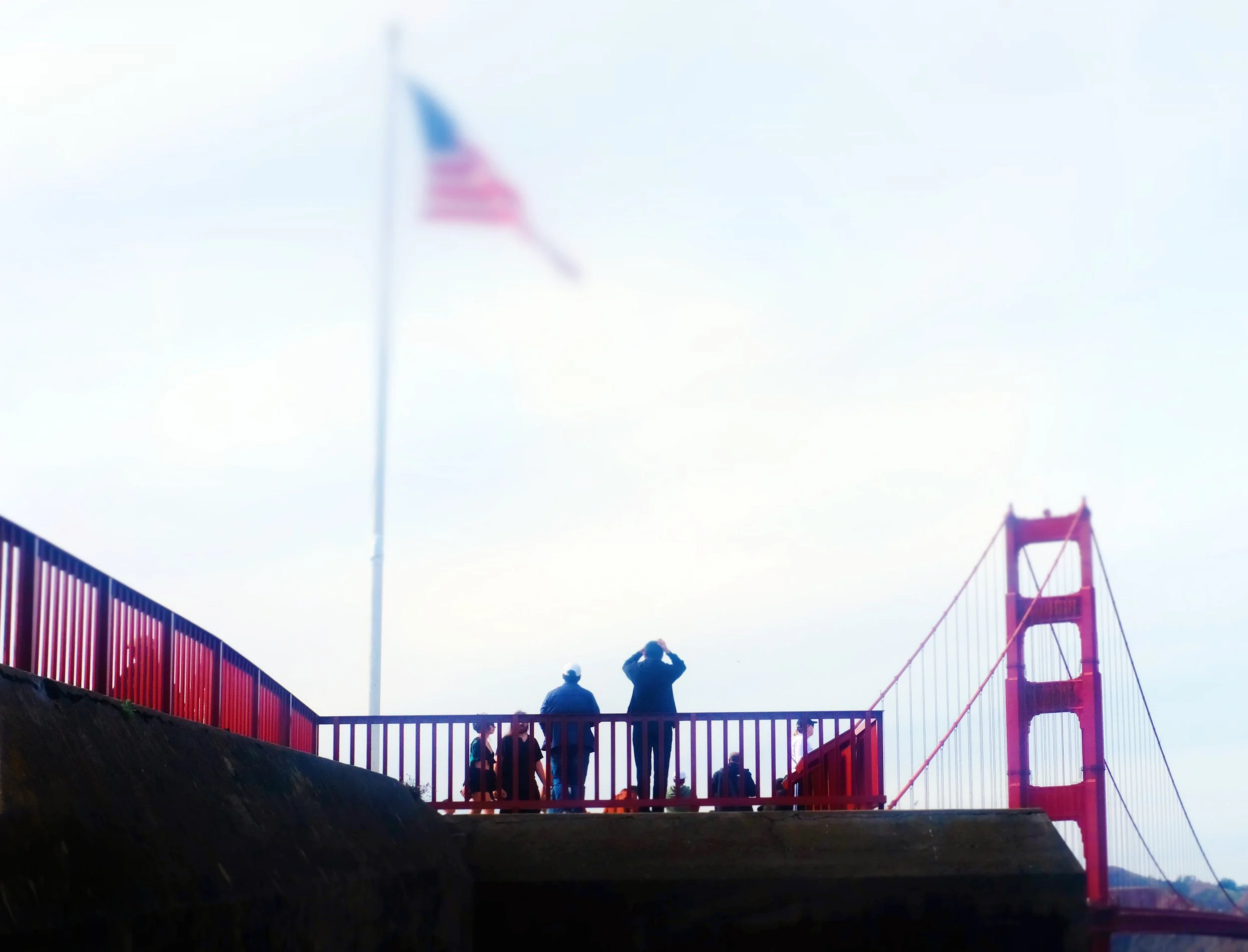 View of the Golden Gate Bridge in San Francisco with people standing on a viewing platform, some taking photos. An American flag is visible in the background.