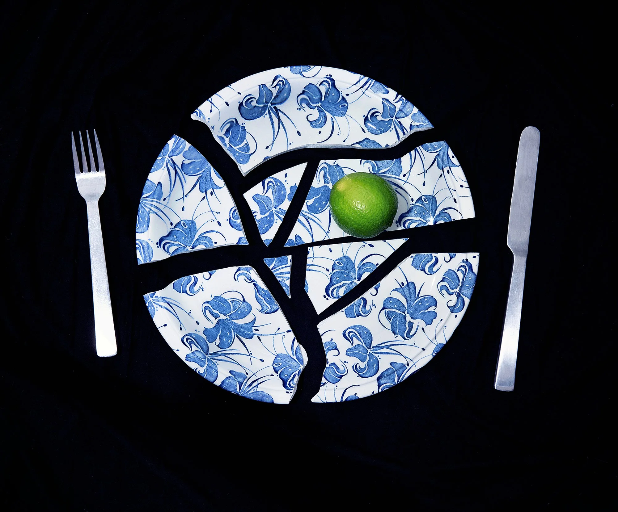 A decorative table setting with a black cutout of a tree on blue and white floral patterned plates, lime, silver fork, and knife against a black background.