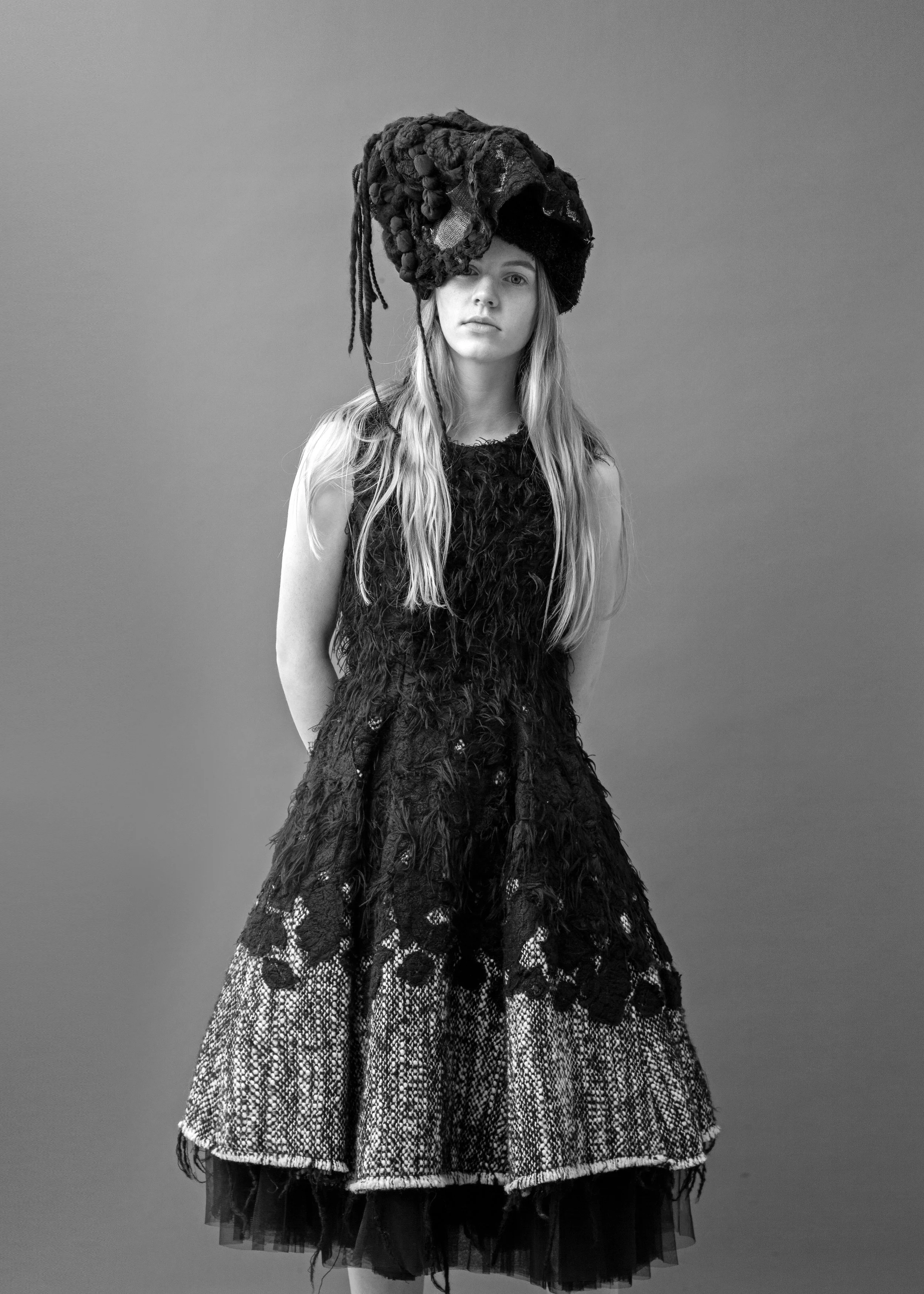 A young woman with long hair wearing an avant-garde dress and headpiece, standing against a plain background.
