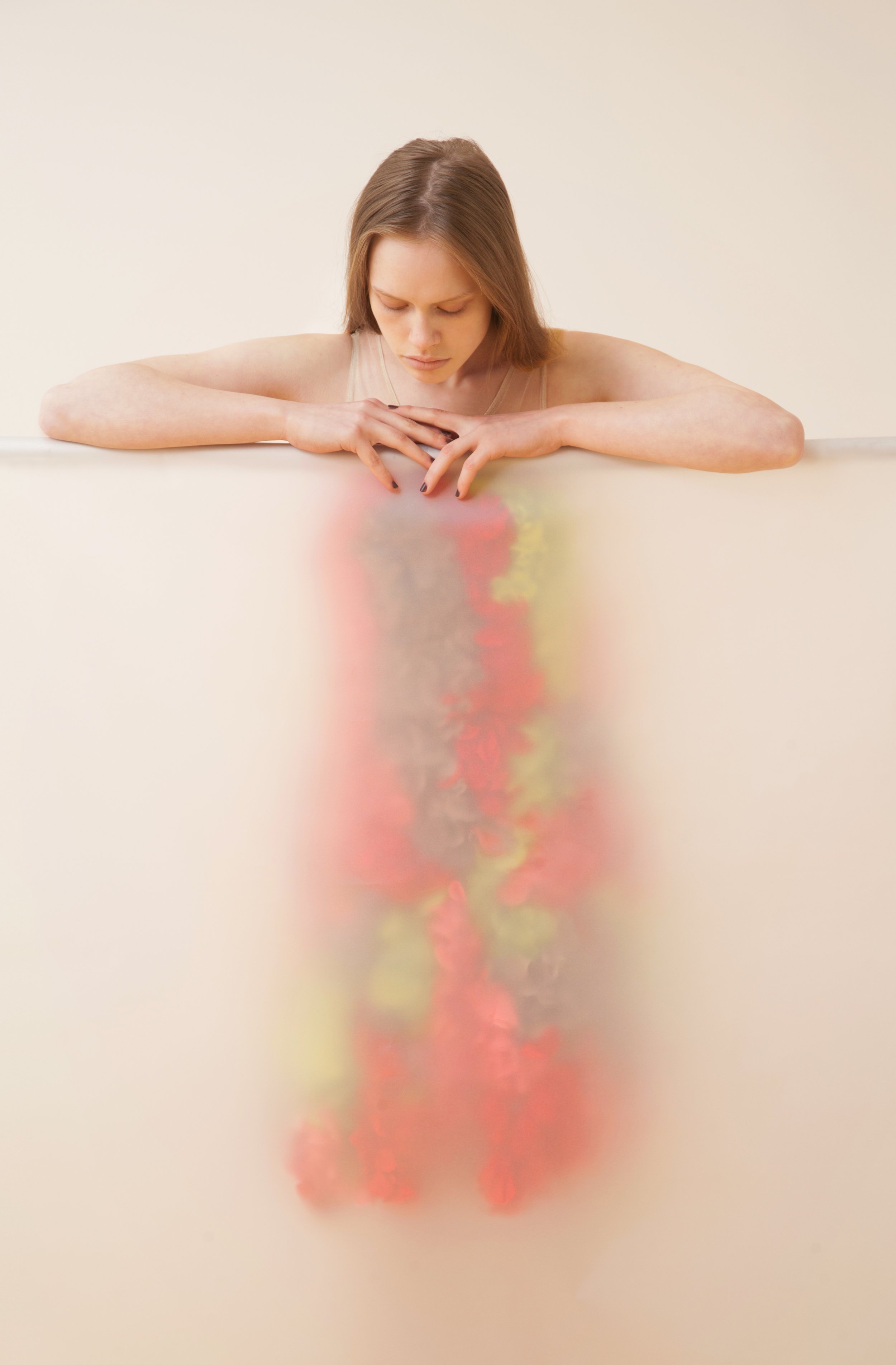 A young woman with short brown hair looking down, resting her arms on a surface, with a colorful and slightly blurred reflection below her.