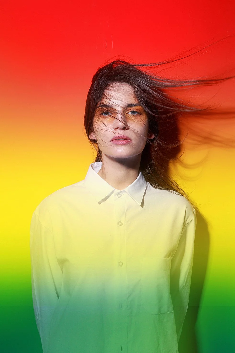 A woman with long dark hair and blue eyes wearing a white button-up shirt against a colorful background blending red, orange, yellow, and green.