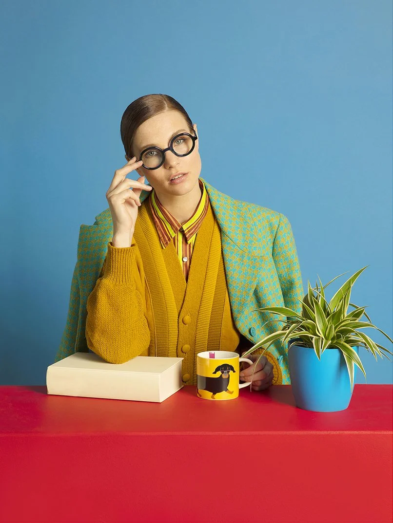 A woman with brown hair pulled back, wearing large round glasses, a yellow cardigan, and a patterned blazer, sitting at a red table with a white book, a coffee mug with a black dog, and a blue pot with a green plant against a solid blue background.