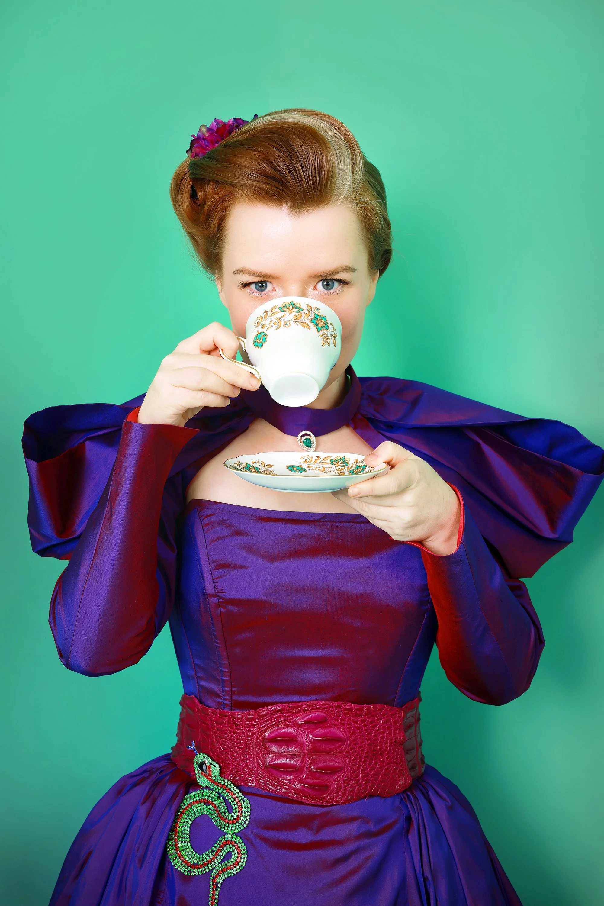 A woman with red hair and blue eyes holding a white and gold-teal teacup and saucer, wearing a purple satin dress, a red textured belt, and a large green and red decorated brooch against a green background.