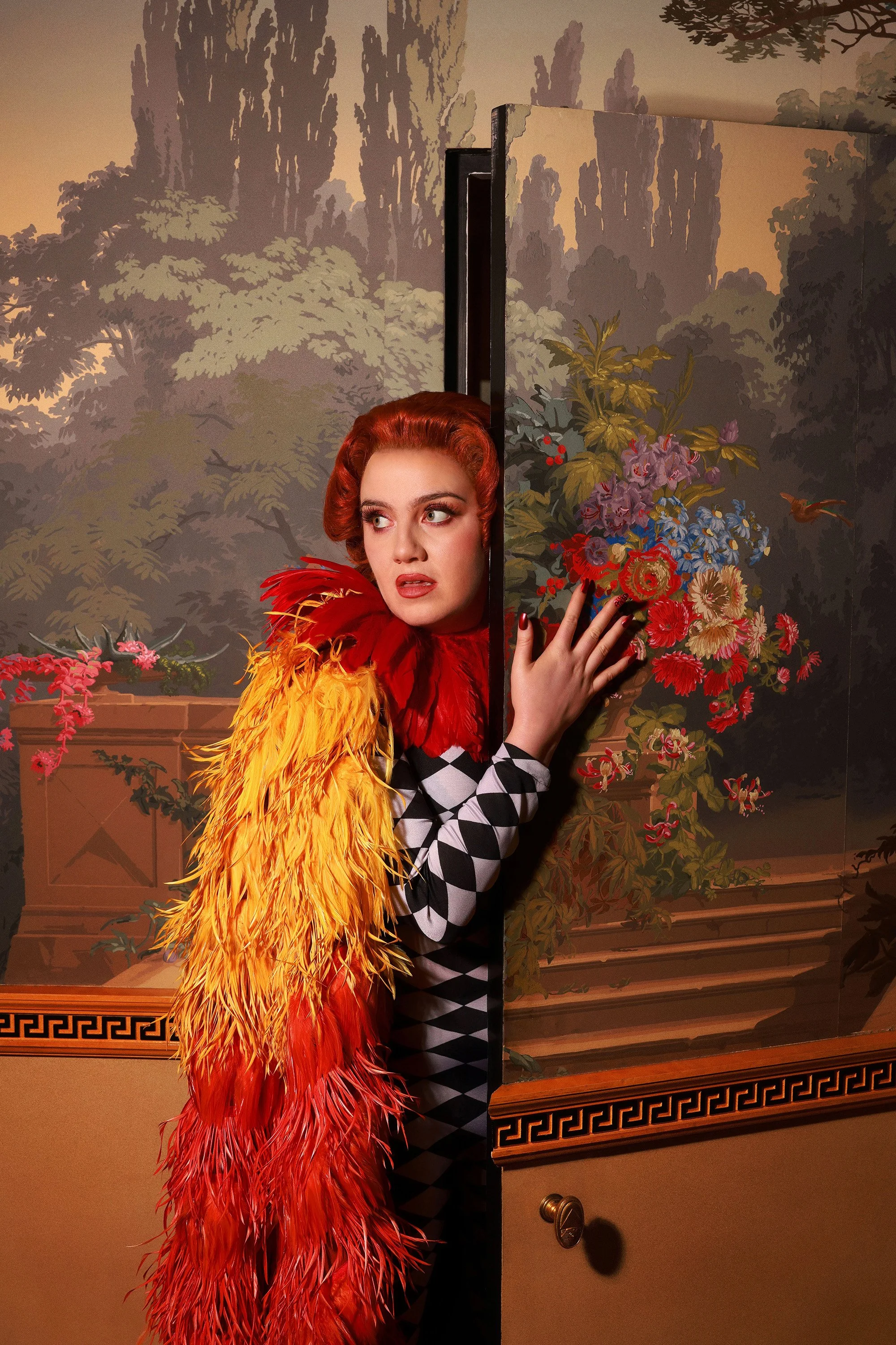 CMAT wearing a red and orange feathered coat, looking to her right, partially hiding behind a black screen with a floral wallpaper background.