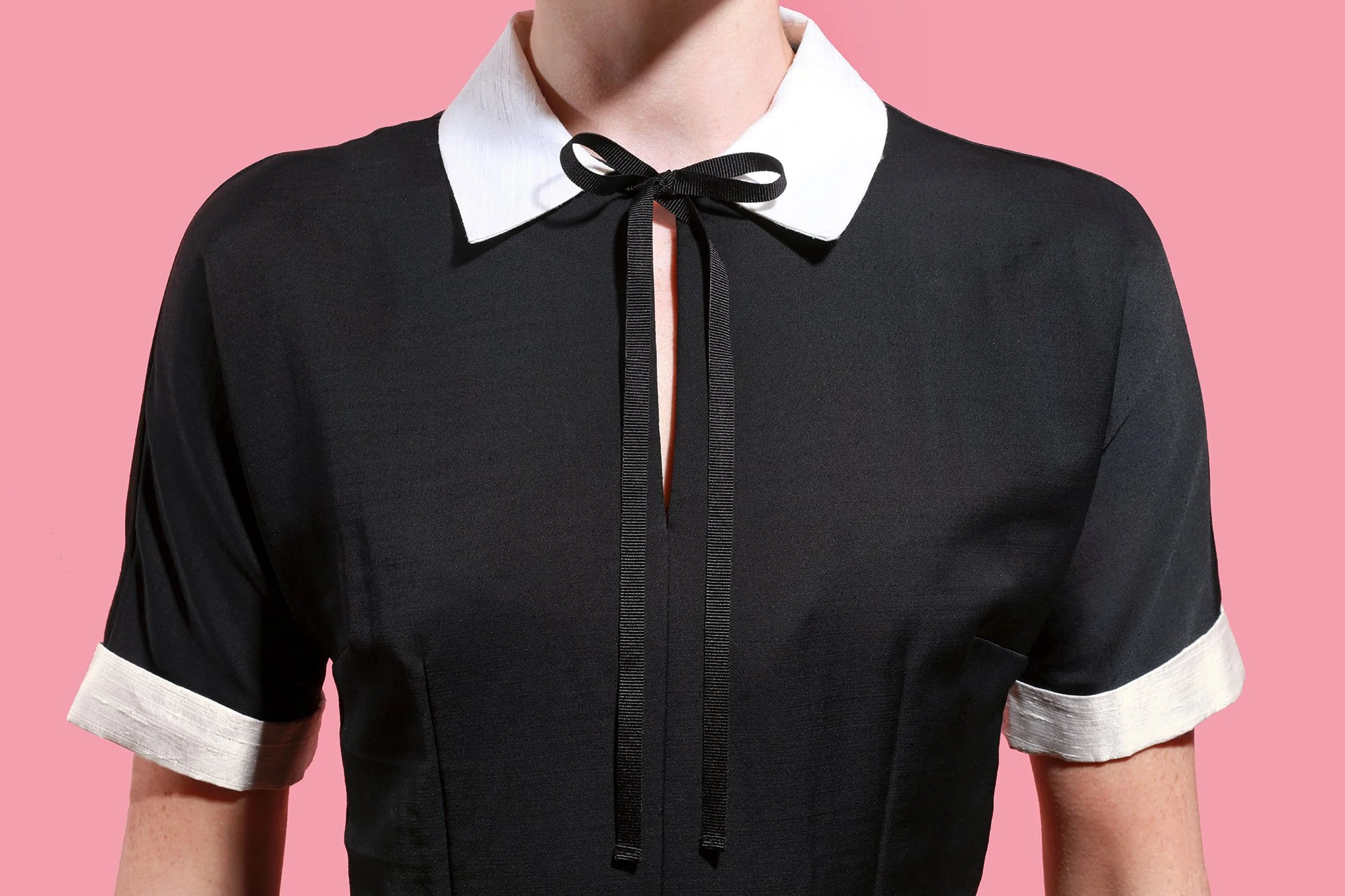 Close-up of a woman's upper body wearing a black dress with a white collar and white cuffs, tied with a black ribbon at the neckline, against a pink background.