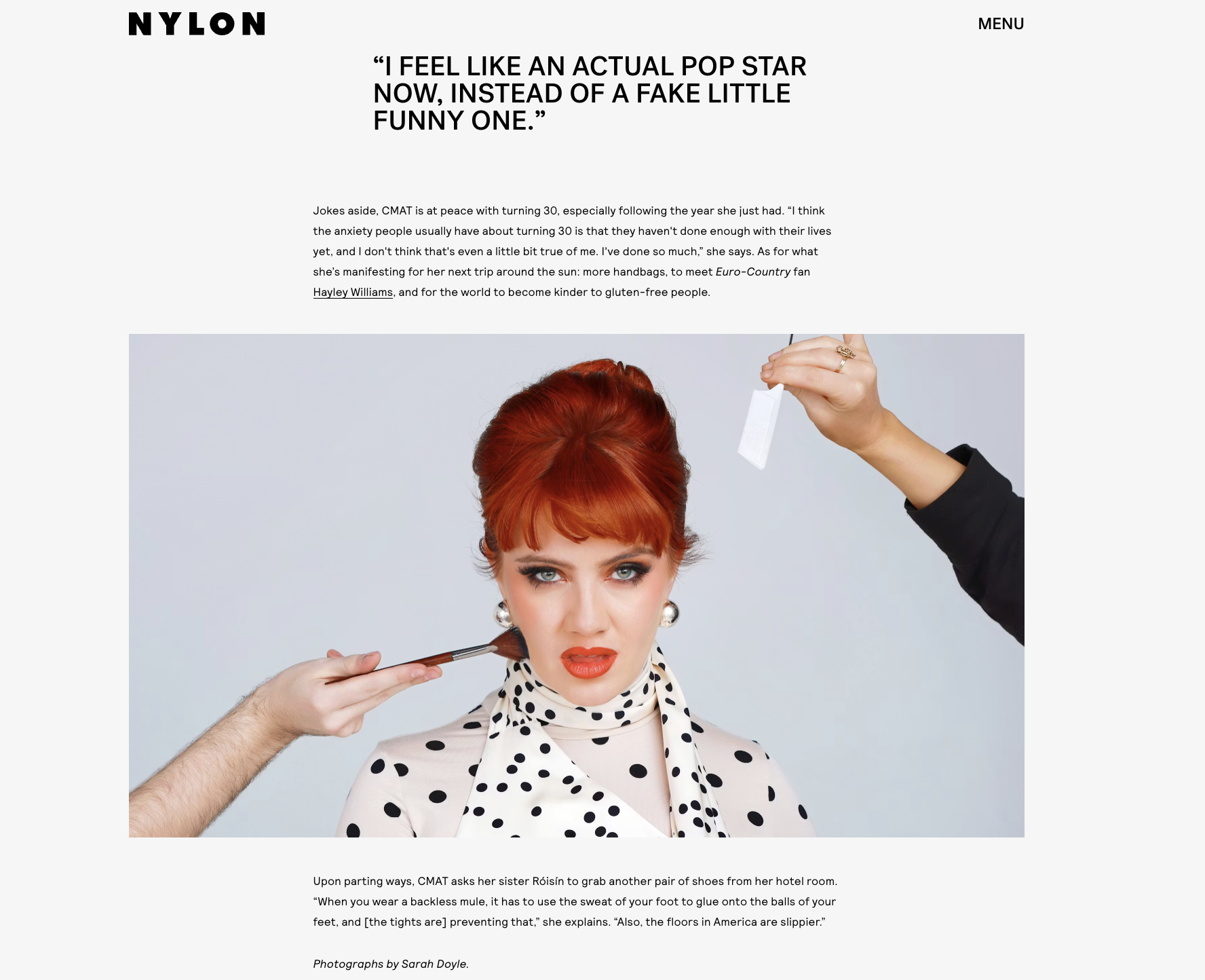 A woman with red hair and vintage makeup having makeup applied by two hands, one in black holding a makeup brush and the other in a black sleeve holding a white object, possibly a makeup sponge or powder puff, against a light blue background.