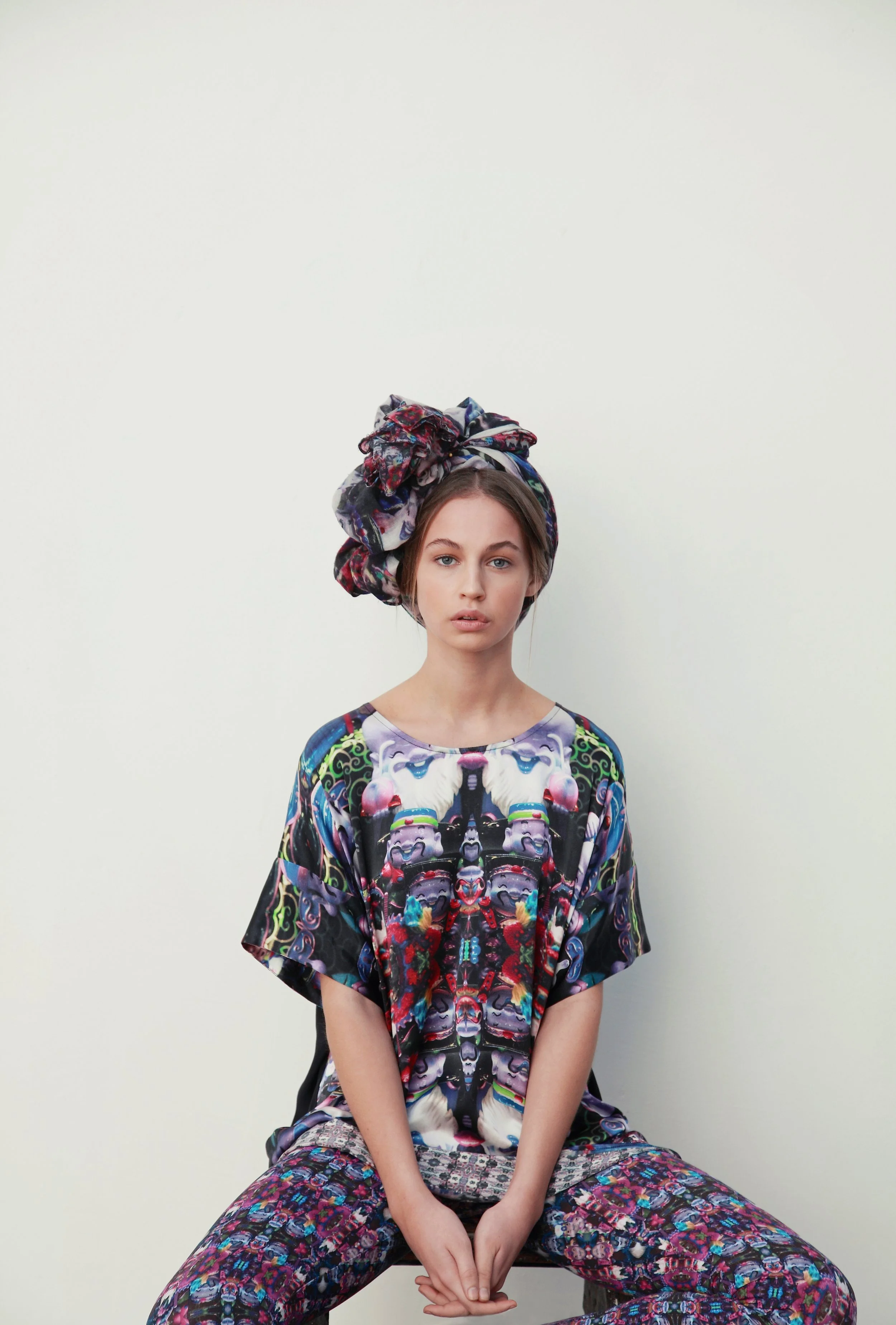 A young woman with light skin and straight brown hair, wearing a colorful, patterned outfit and headscarf, sitting cross-legged against a plain, light-colored background.