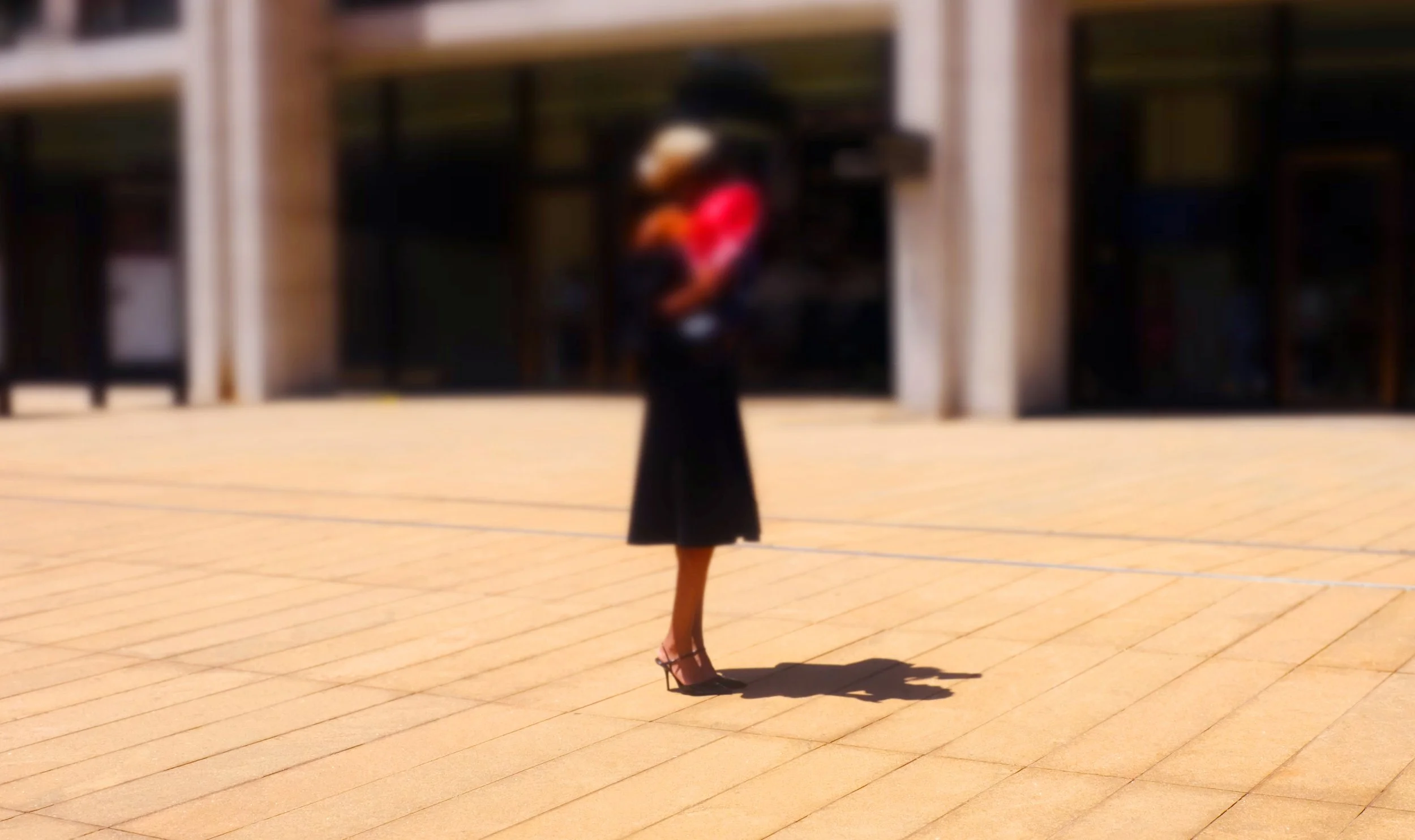 A woman in a black dress and high heels walking outdoors on a sunny day, casting a shadow on the paved ground, with a building in the background.