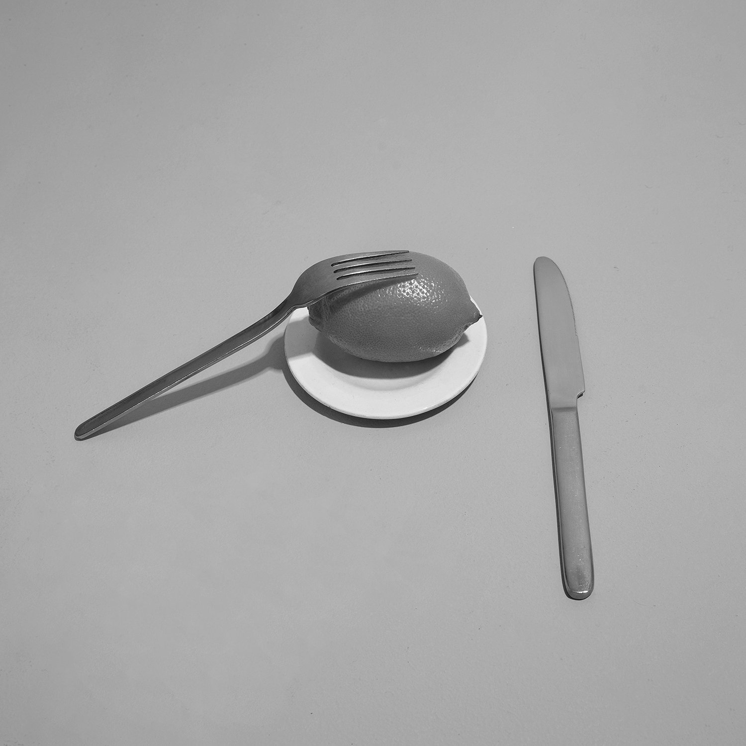 A lemon on a small white plate, with a fork resting on top, and a knife placed on the table beside it, all in black and white.