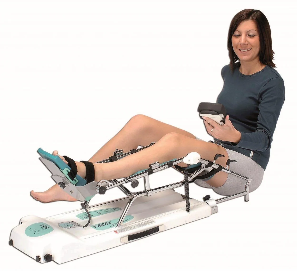 A woman sitting on a medical knee rehabilitation machine, holding a remote control, smiling, with her legs extended and secured by straps, in a clinical setting with a plain white background.