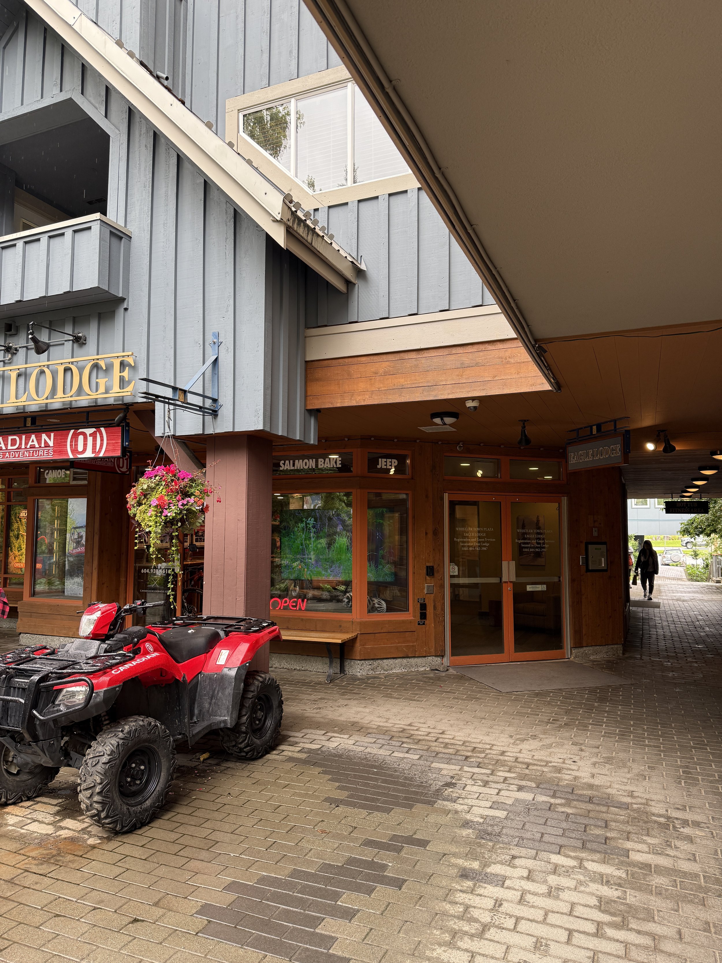 Exterior view of a restaurant with wooden and blue-gray paneling, a hanging flower basket, an ATV, and a person walking in the background.