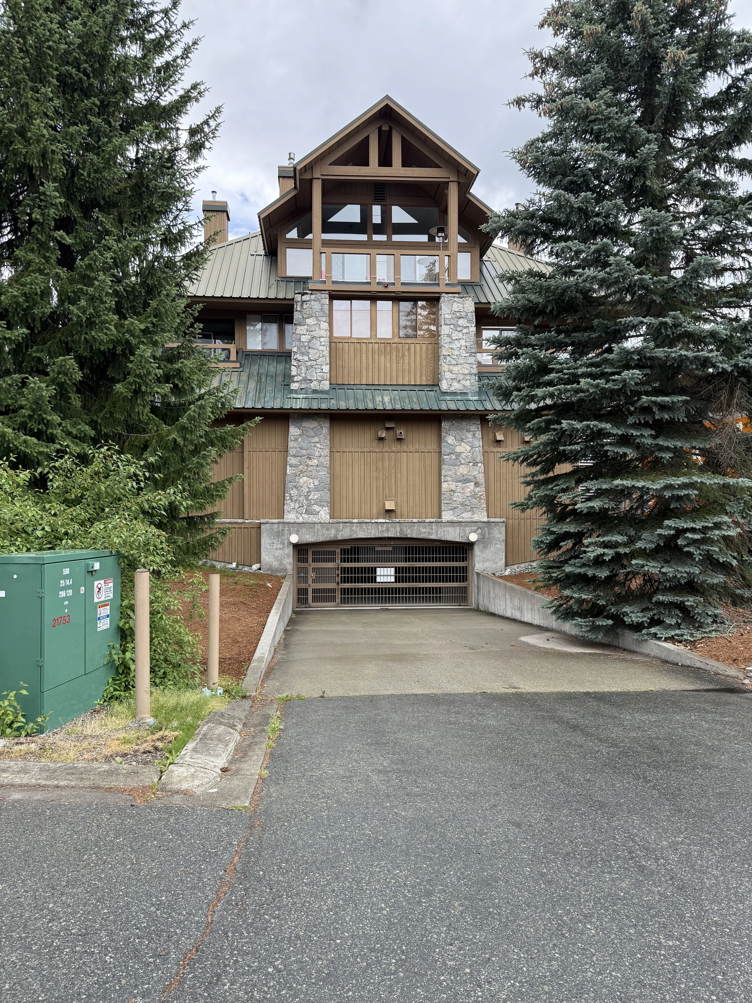 Front view of a multi-story house with a combination of wood and stone exterior, surrounded by trees, with a driveway leading to a garage.