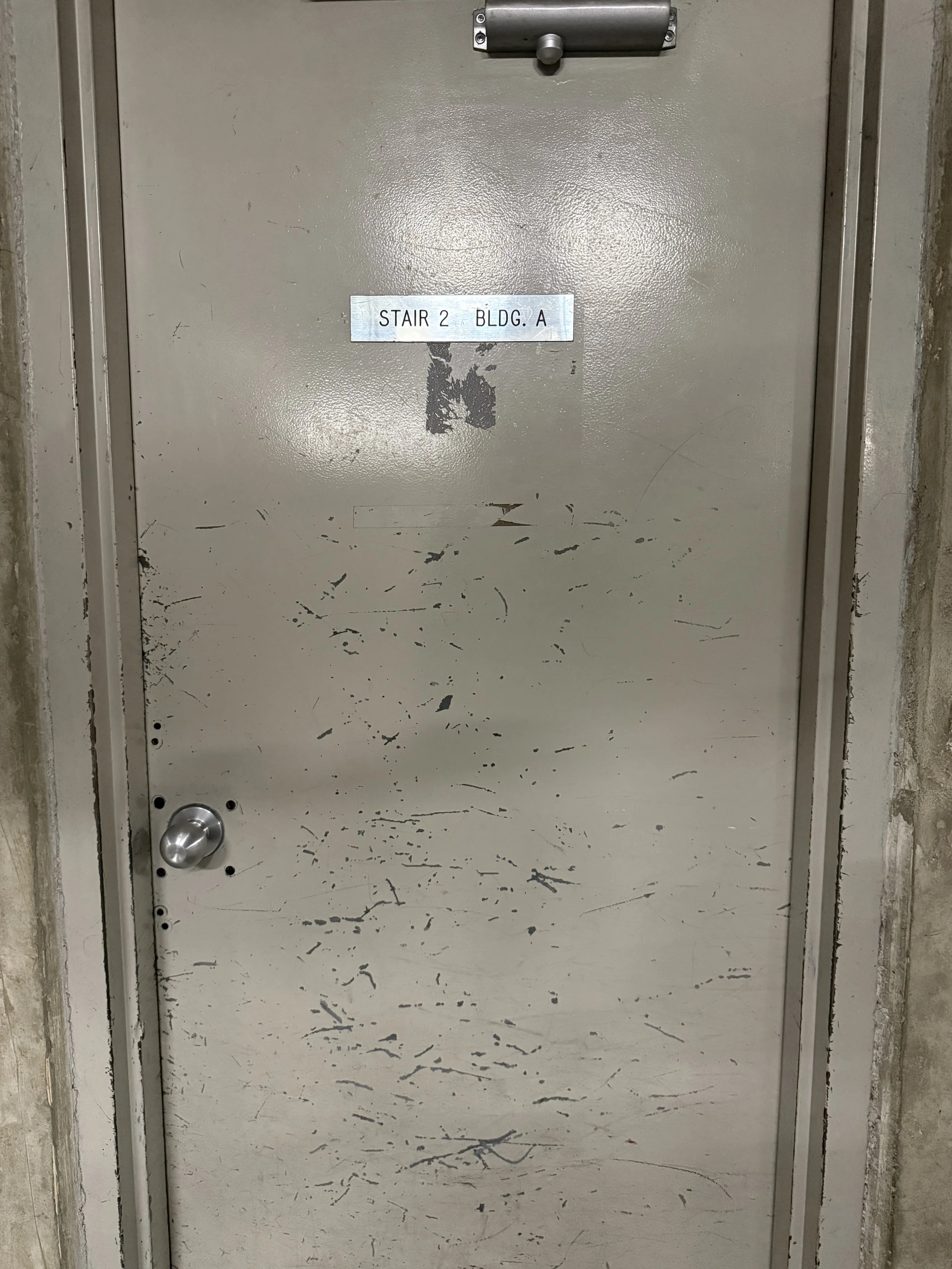 A metal door with a handle on the left side, a latch at the top, and a sign that reads 'STAIR 2 BLDG. A' attached in the upper middle part of the door. The door has scratches and marks, and is set within a concrete wall.