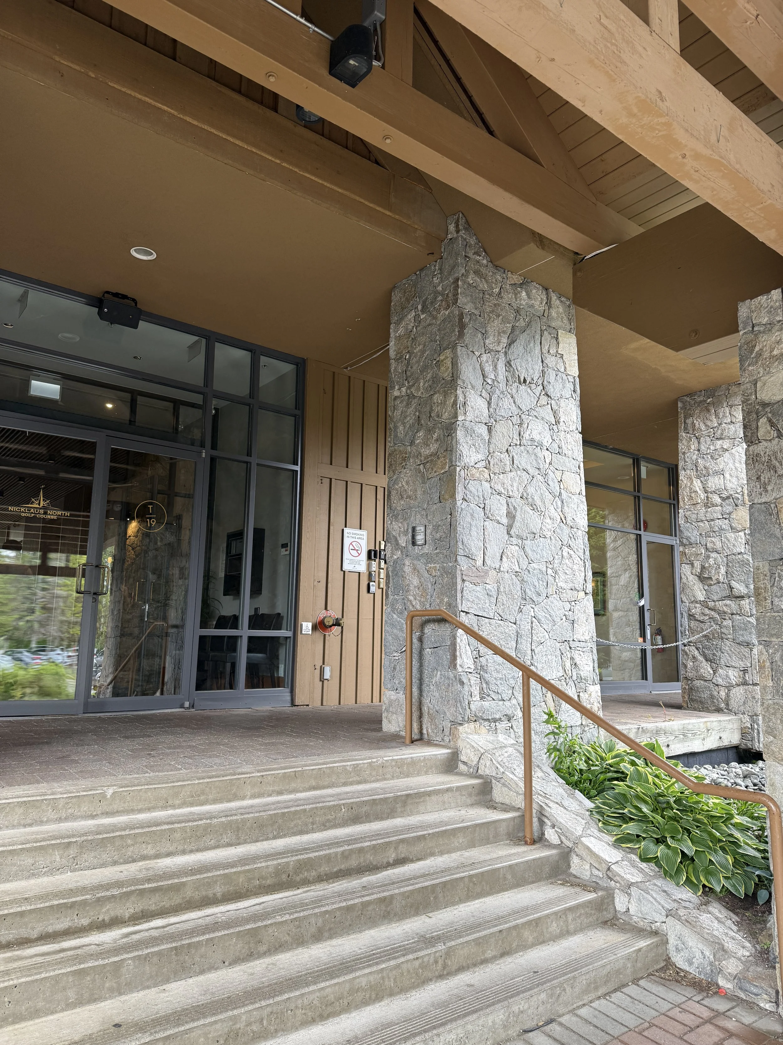 Exterior view of a building with stone and wood walls, glass door, and concrete stairs, with a metal handrail and green plants beside the stairs.