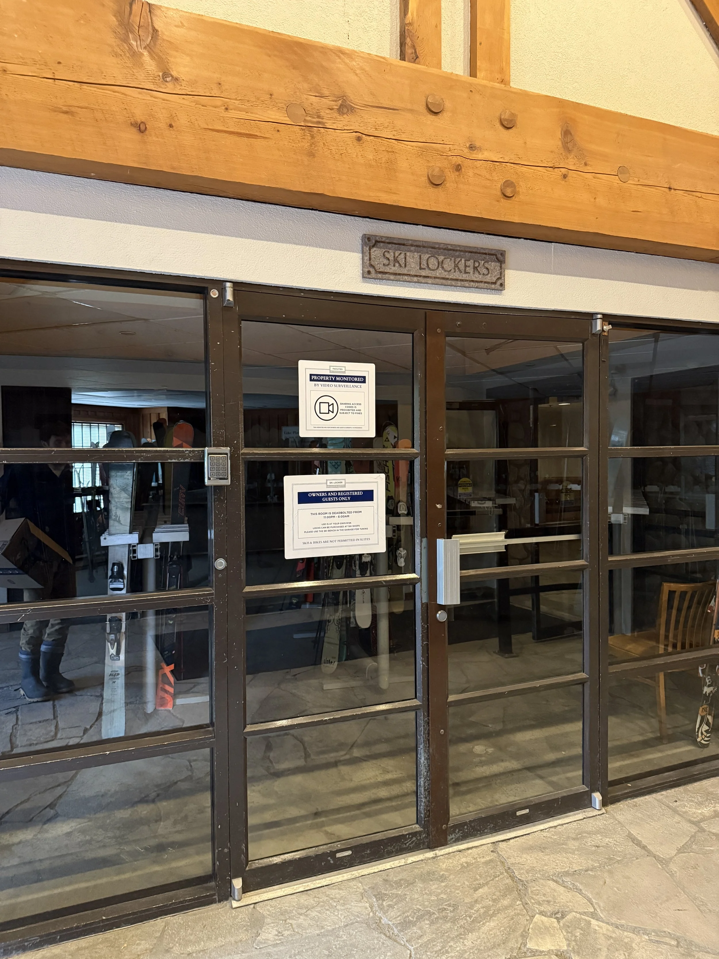 Glass door with signs, leading to ski lockers, inside a building with stone floor and wooden trim.