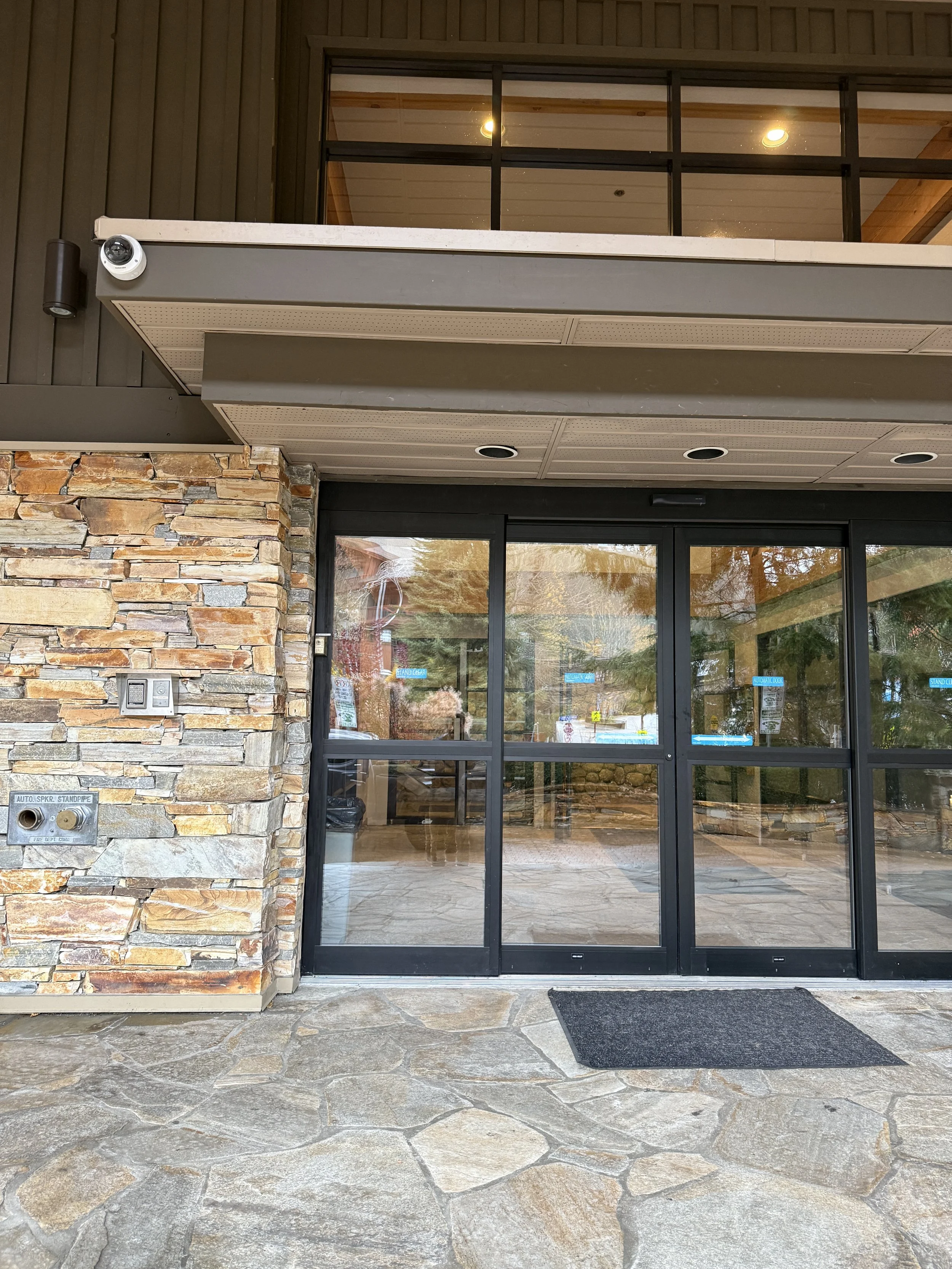 View of the entrance to a building with glass double doors, stone wall on the left, and a black welcome mat on a stone patio. The upper level has a balcony with a glass railing and interior lighting visible through windows.