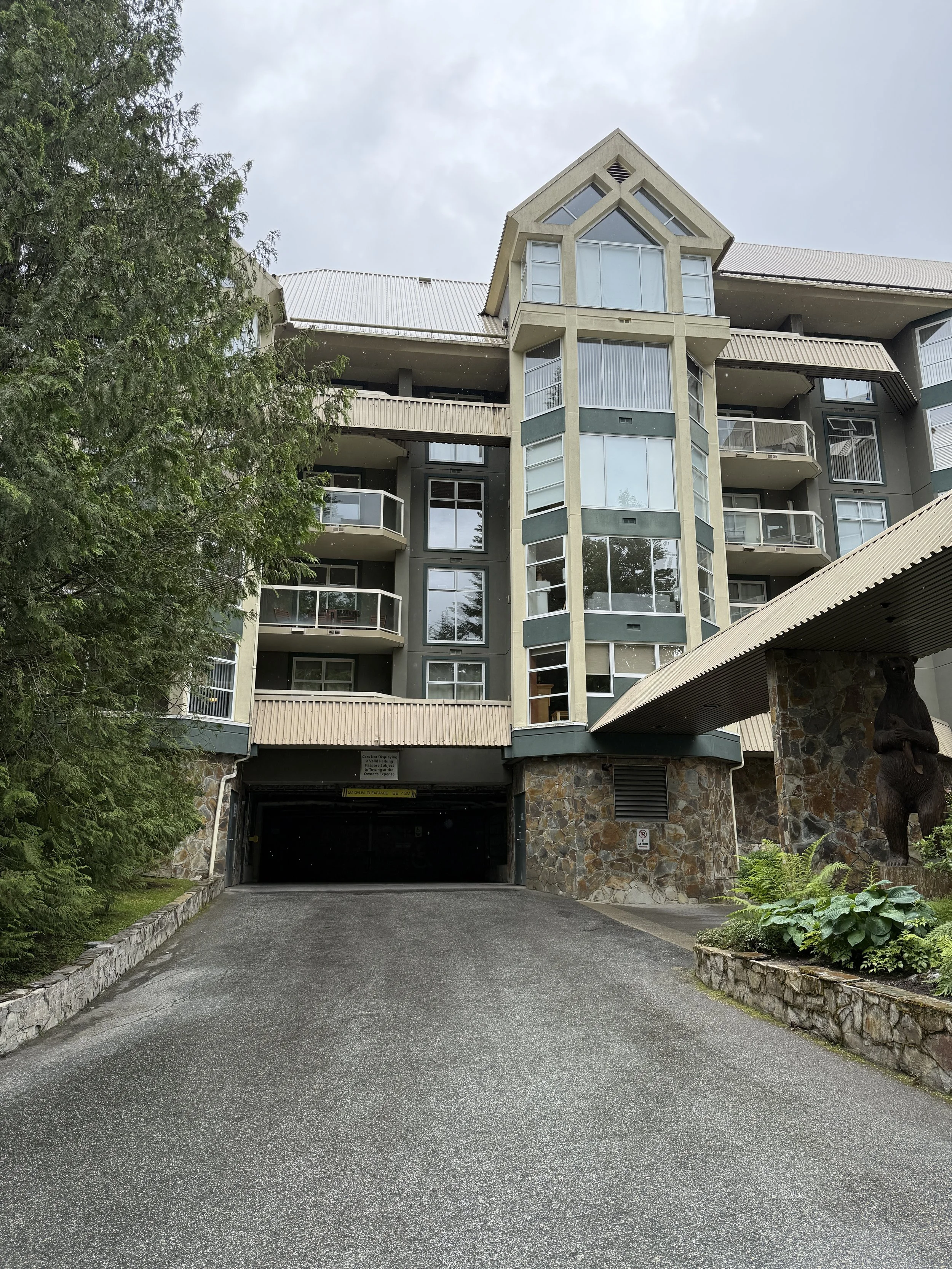 Multi-story residential building with a stone and glass exterior, balconies, and a sloped driveway leading to a garage at the front, surrounded by greenery, in cloudy weather.