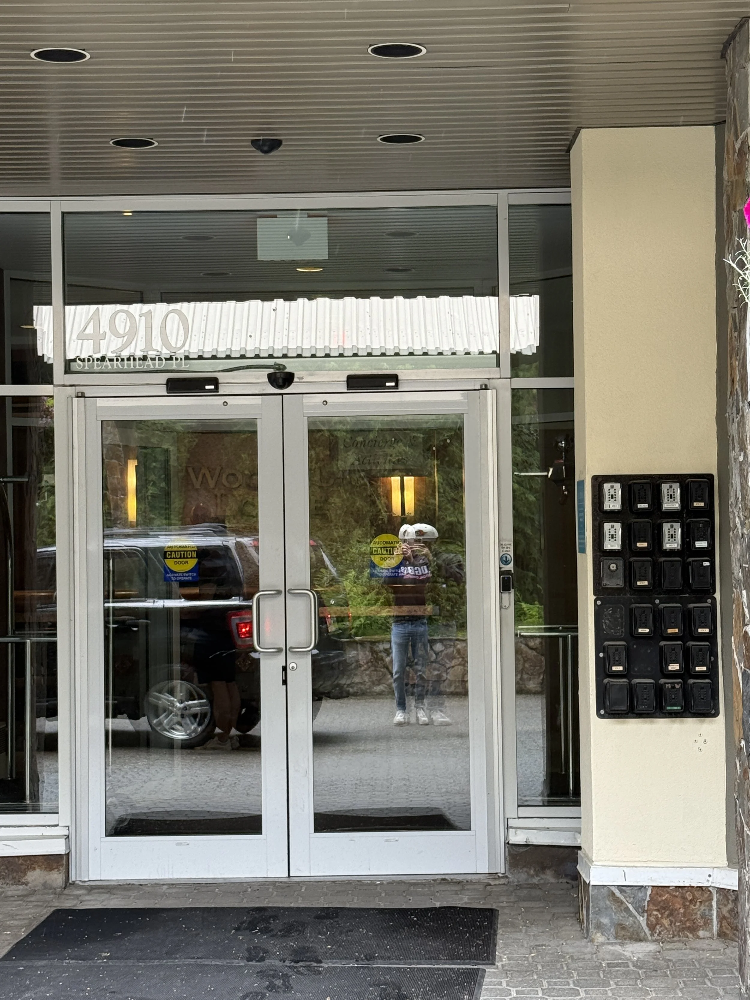 The entrance of a building with a glass door, the address '4910 SPEARHEAD PL' is visible above the door, and there are multiple black mailboxes on the right side. The reflection in the glass shows a person taking a photo, parked cars, and greenery outside.