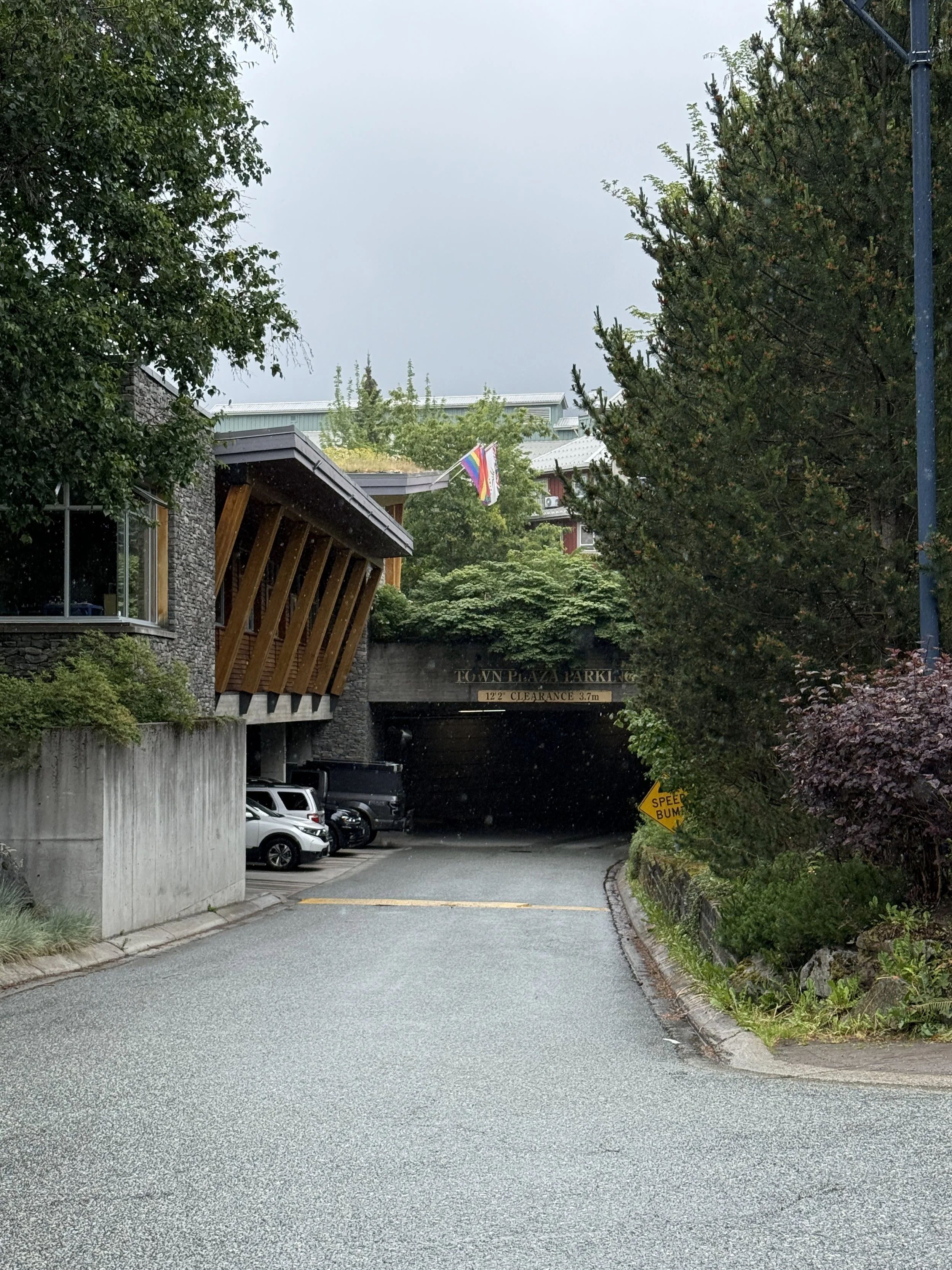 A street leading to a parking garage entrance under an overcast sky, with trees and parked cars on each side and a rainbow flag hanging above the entrance.