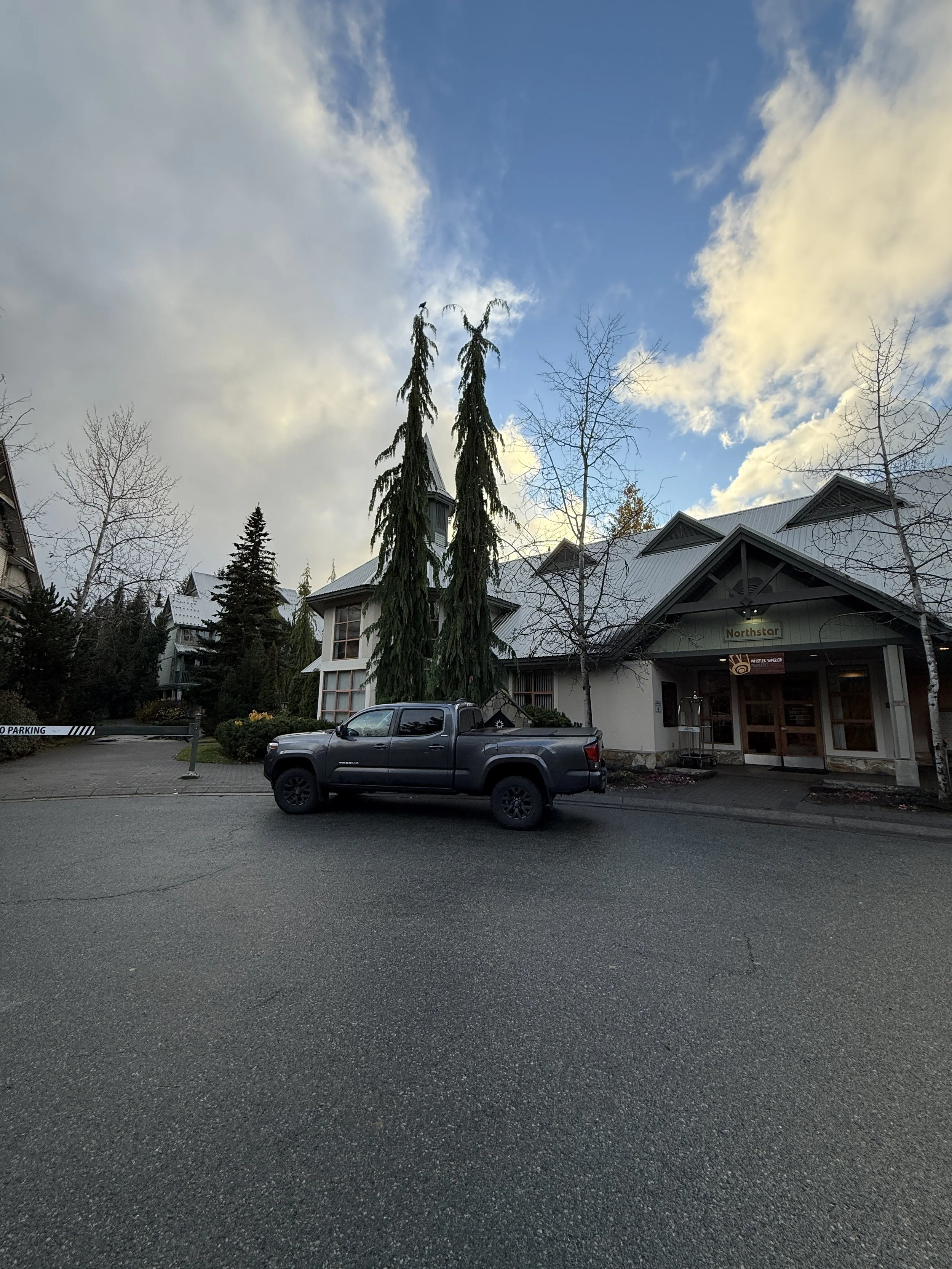 A parking lot in front of a building with a sign that reads 'Northstar,' a gray pickup truck parked, leafless trees, some evergreens, a partly cloudy sky.