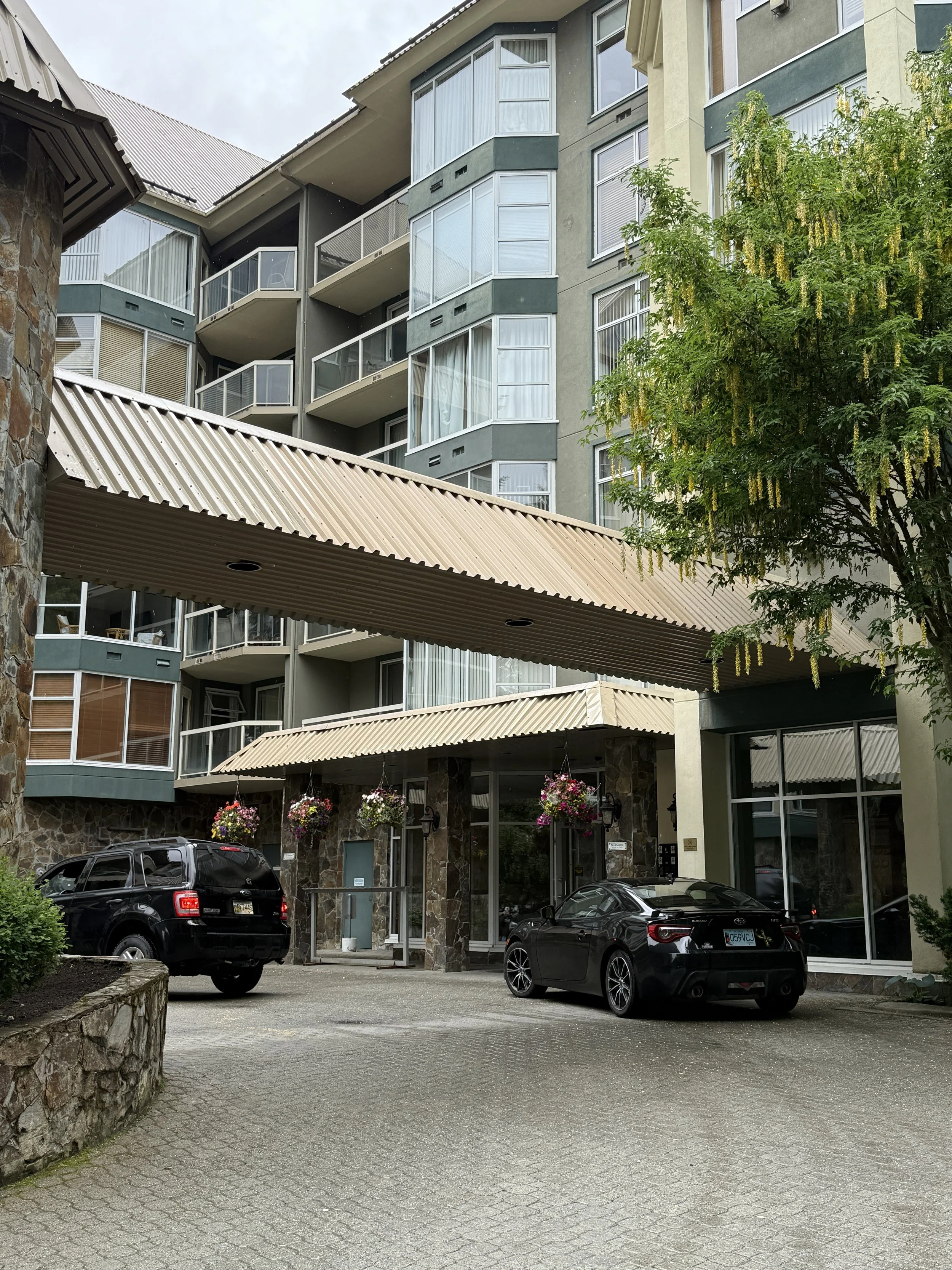 Multi-story apartment building with balconies, an entrance canopy, parked cars, hanging flower baskets, and a tree with hanging yellow flowers.
