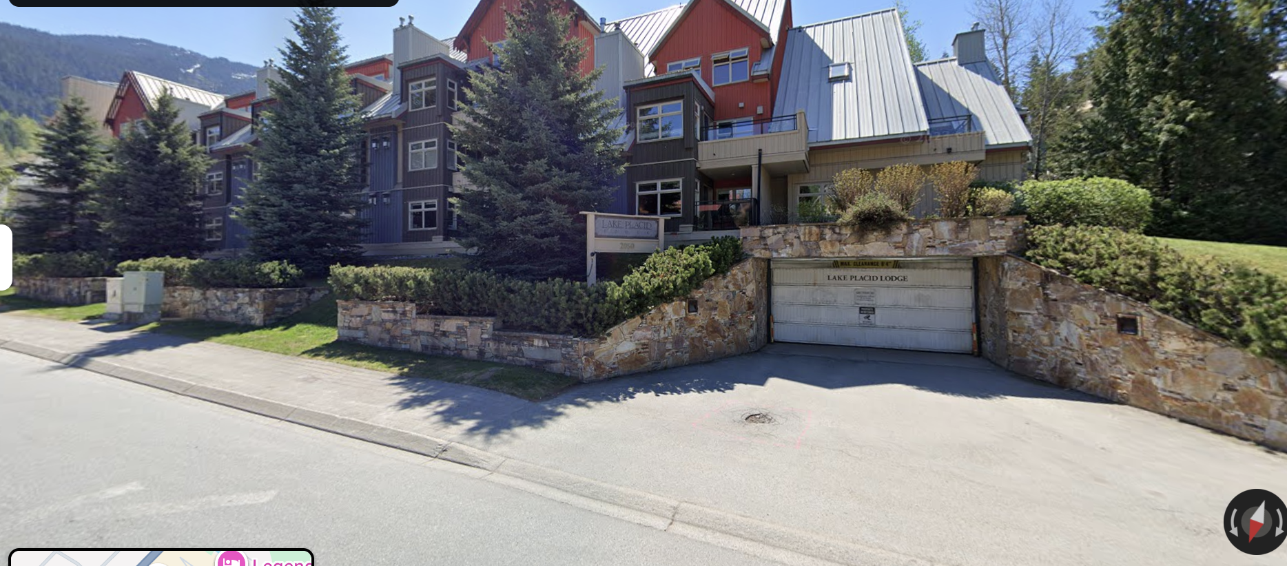 A multi-story residential building with a stone facade at the base, surrounded by tall evergreen trees and landscaped bushes. The building has rustic elements with a combination of grey and red exterior walls, metal roofs, and large windows. A sign reads "Lake Placid" near an underground garage entrance.