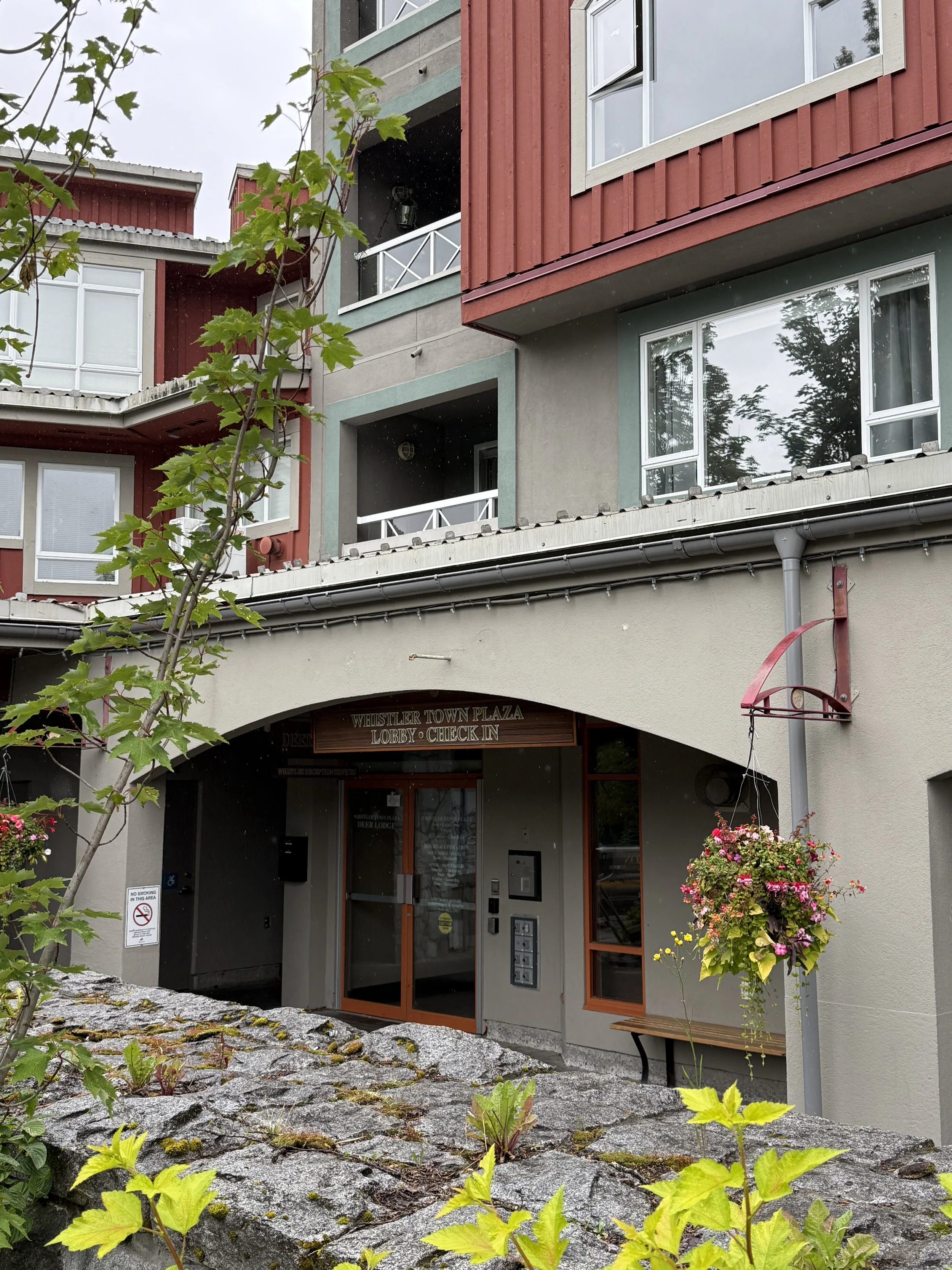 The image shows the entrance to a building with a sign that reads 'Whistler Town Plaza Lobby Check In.' There are gray and red architectural elements, a small tree in the foreground, and hanging flower baskets on the right side.