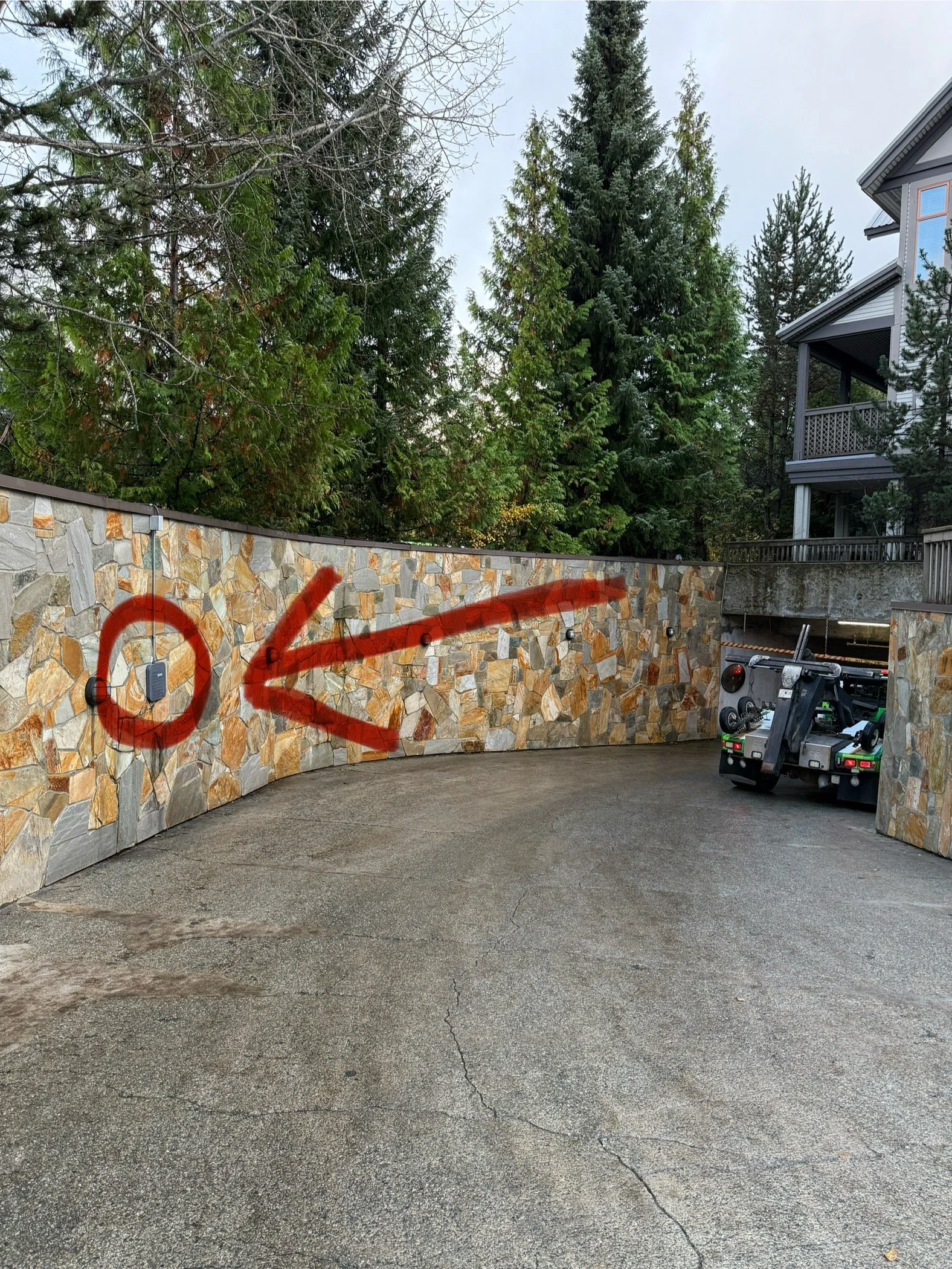 A paved driveway with a stone wall on the left side, marked with a red arrow and circle drawn with spray paint. The wall has small security cameras or lights mounted on it. Behind the wall are tall evergreen trees, and a multi-story house with a porch is on the right side. There is a vehicle with equipment parked near the driveway.