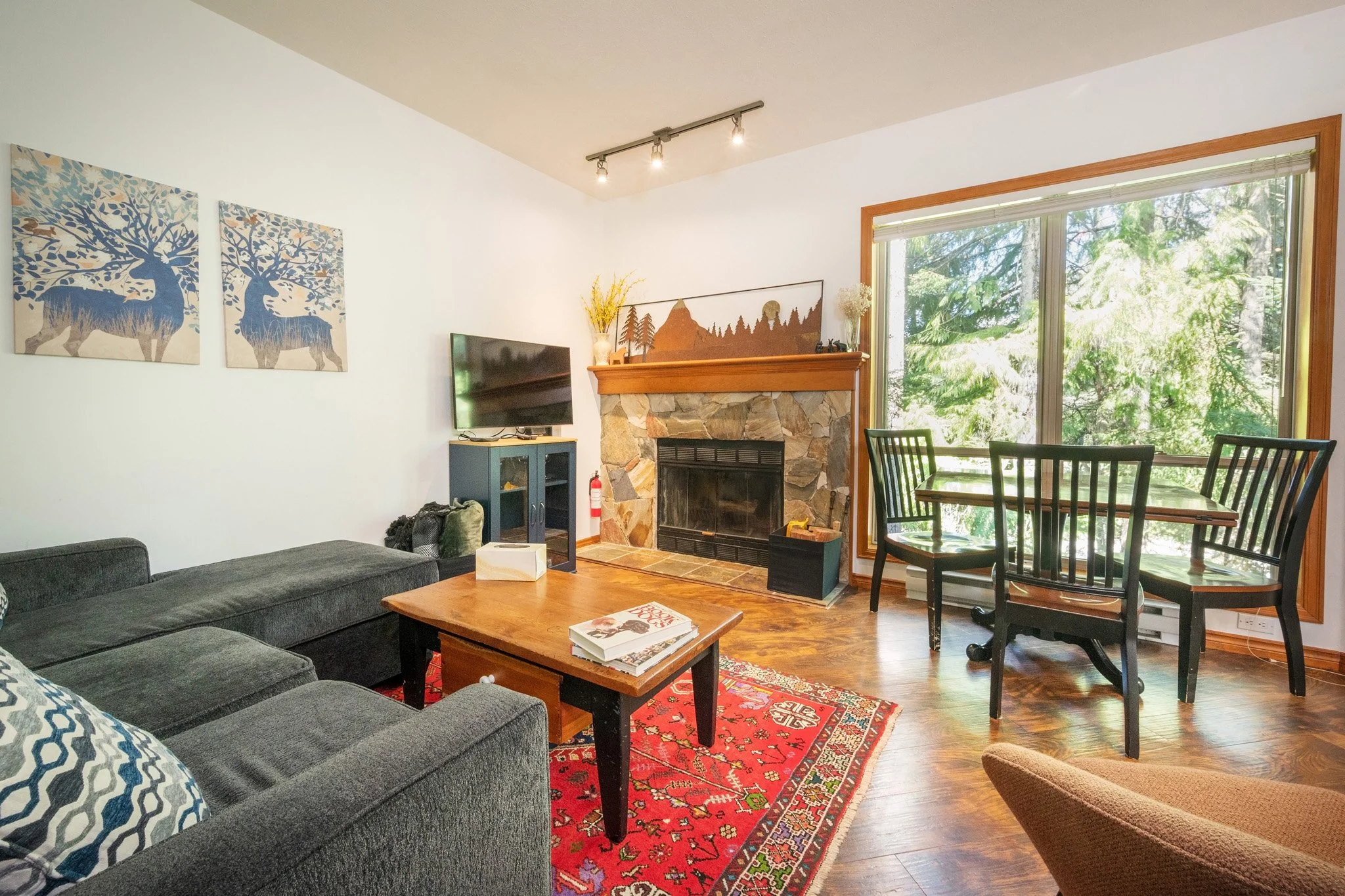Living room with a gray sectional sofa, wooden coffee table with books, fireplace with stone surround, large window showing trees outside, dining table with black chairs, wall art of deer and trees, and ceiling track lights.