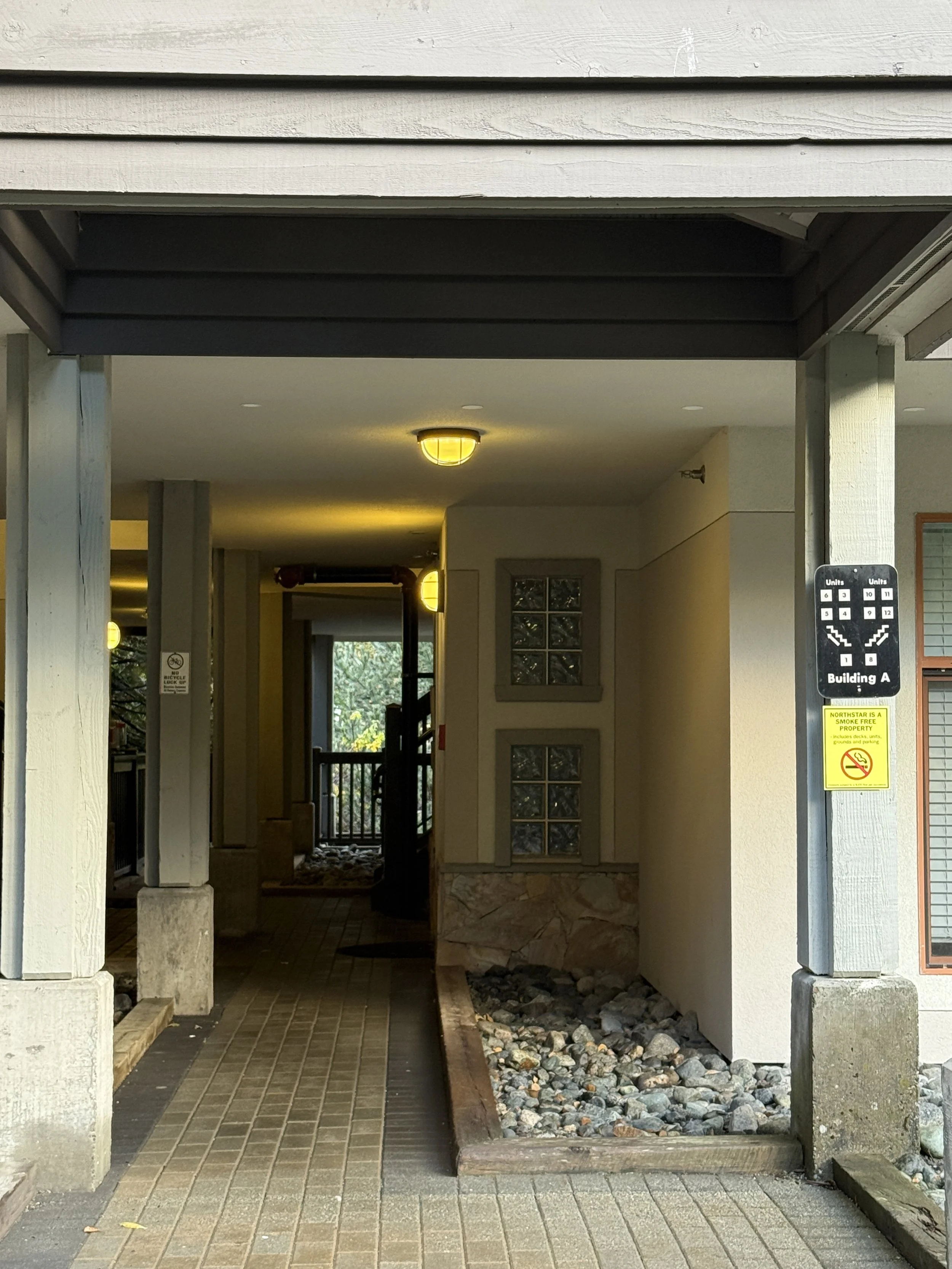 Exterior view of a building entrance with a covered walkway, light yellow walls, glass block windows, and decorative rocks in a landscaped bed.